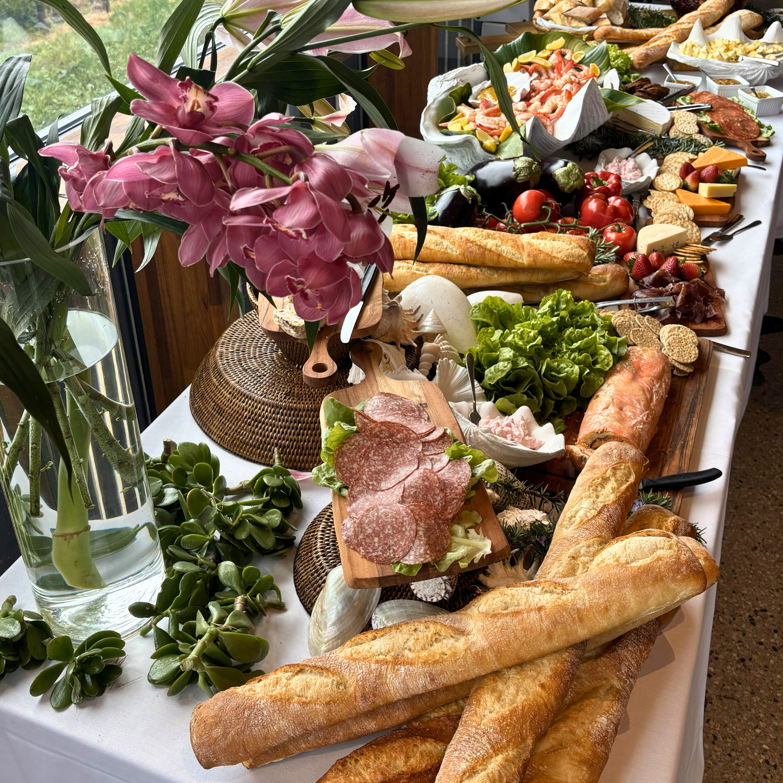 A buffet table with assorted bread, charcuterie, cheeses, vegetables, and fresh fruit, decorated with pink orchids and green plants.