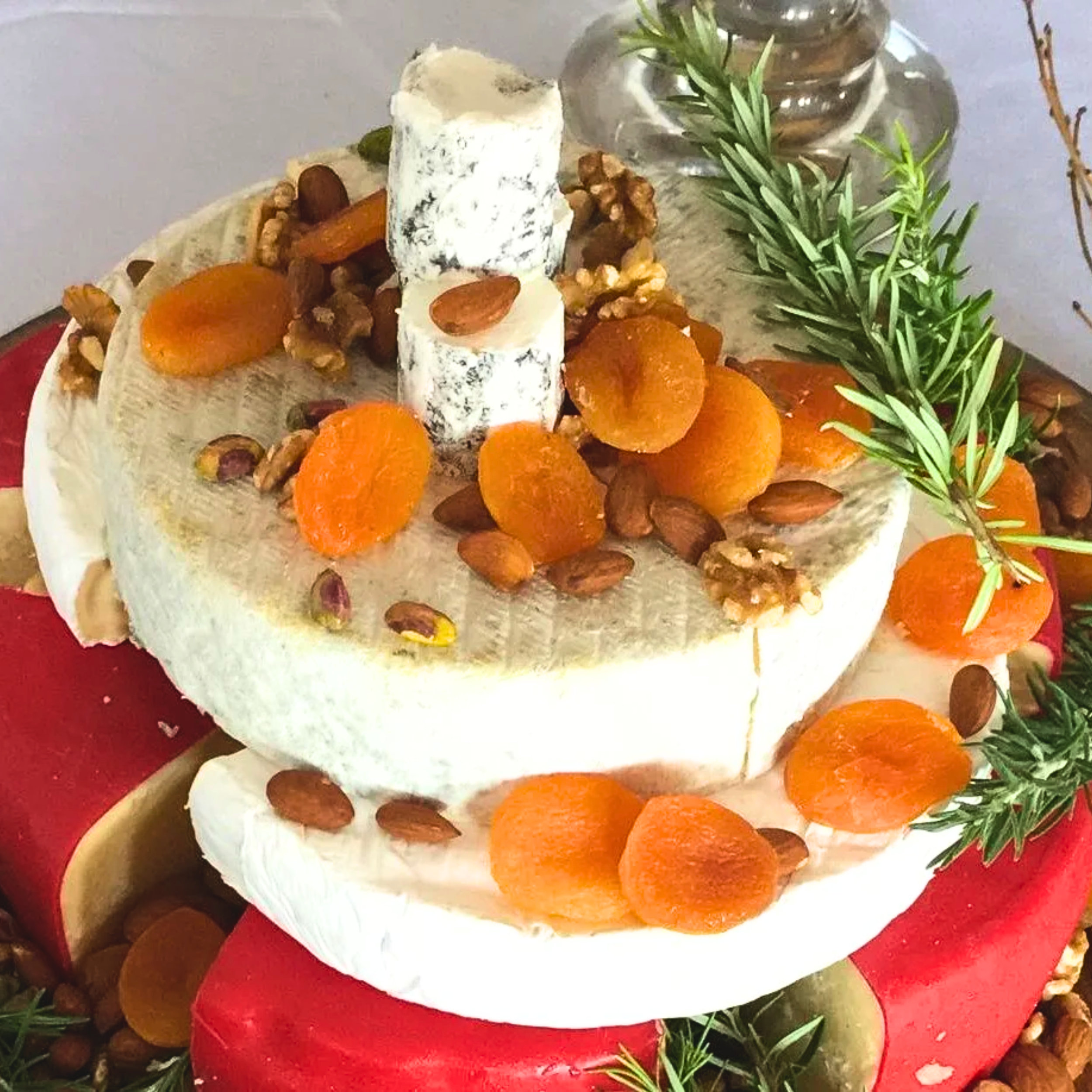 A cheese wheel decorated with dried apricots, walnuts, pistachios, and a sprig of rosemary.