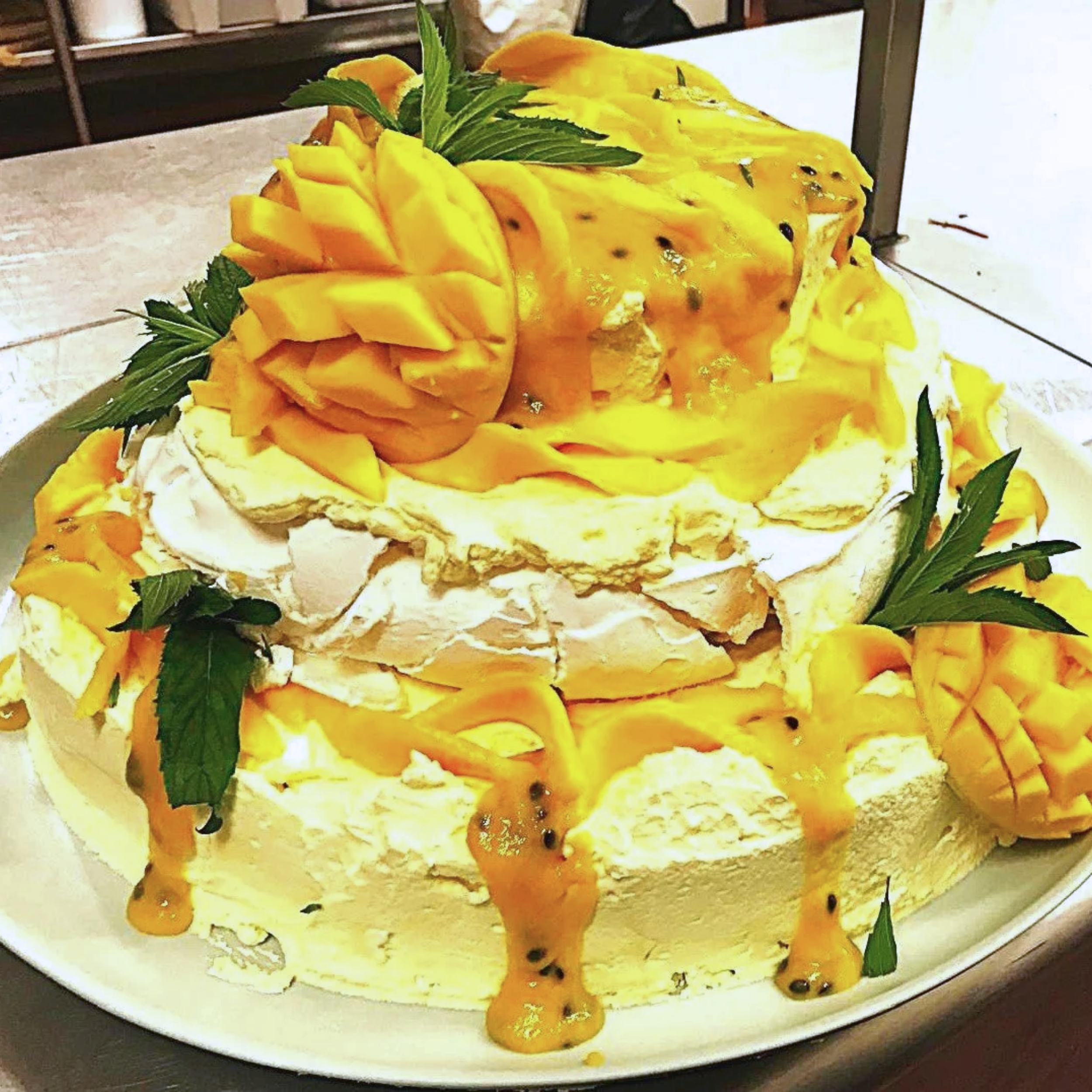 A pavlova cake decorated with sliced mango and green leaves, topped with a carved mango flower and passionfruit sauce.
