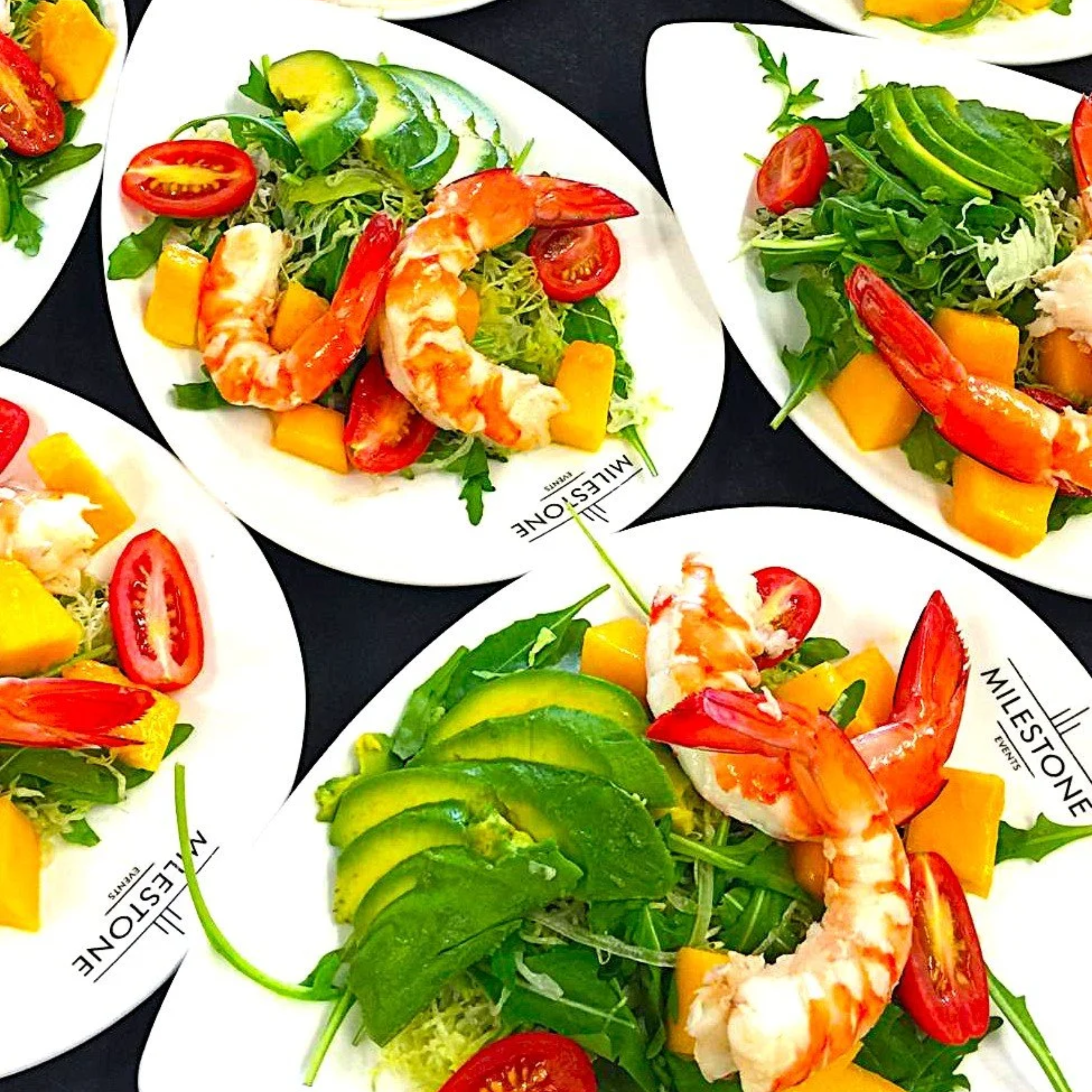 Salad with cherry tomatoes, mango chunks, avocado slices, lettuce, and cooked shrimp on white oval plates with black surface underneath.