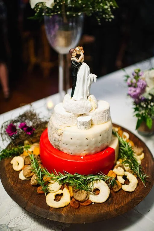 Wedding cake made of large cheese rounds with a bride and groom figurine on top, decorated with nuts and herbs, surrounded by flowers.