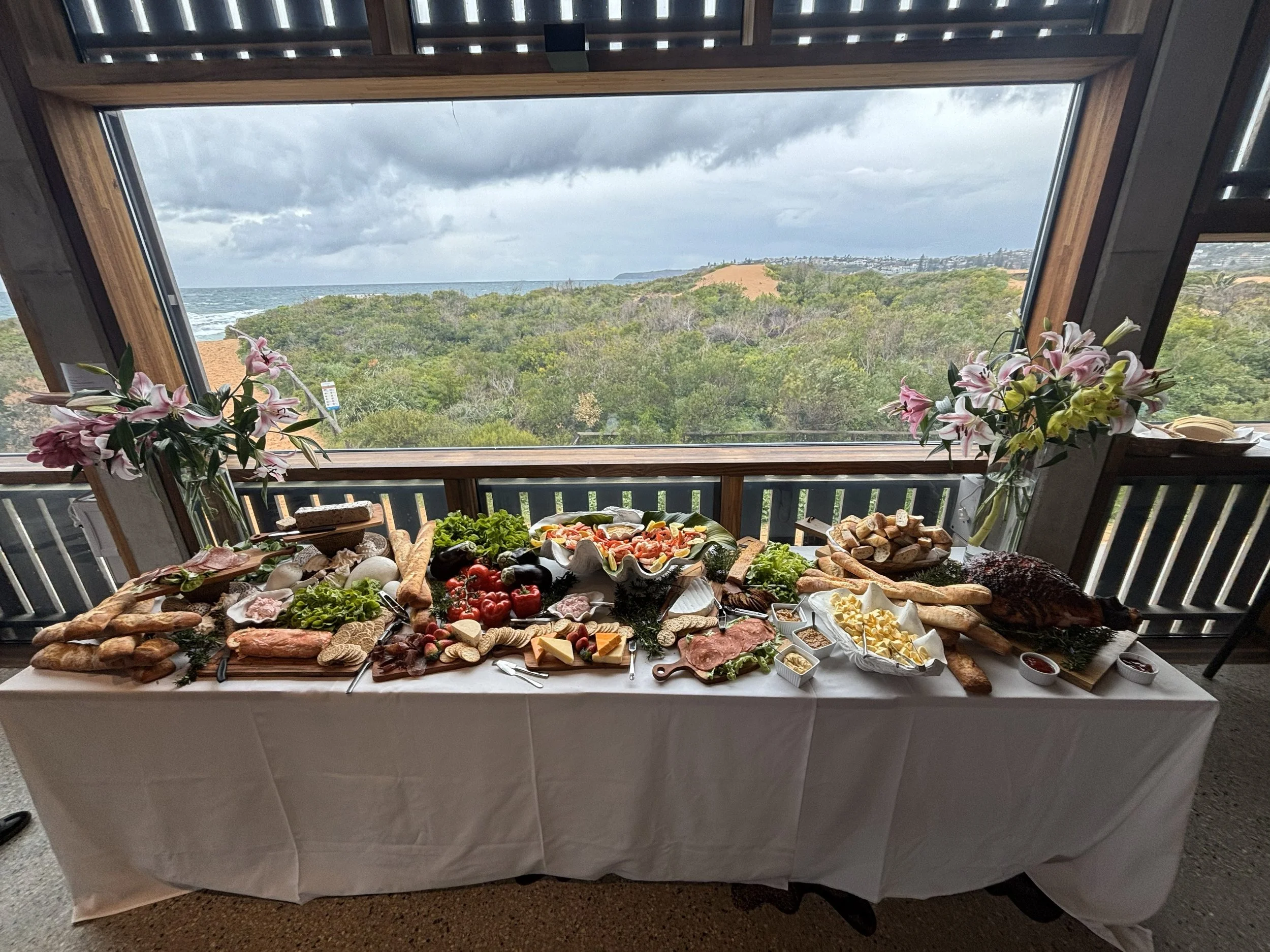 A large buffet table with various foods including bread, cheeses, meats, vegetables, and fruit, set near a window overlooking a scenic landscape with trees, hills, and the ocean under a cloudy sky.