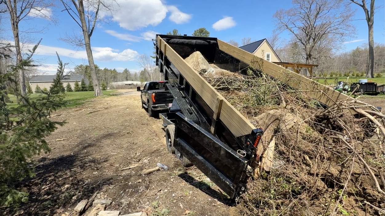 A black dump truck unloading dirt and debris onto a dirt lot with scattered branches and trees nearby.