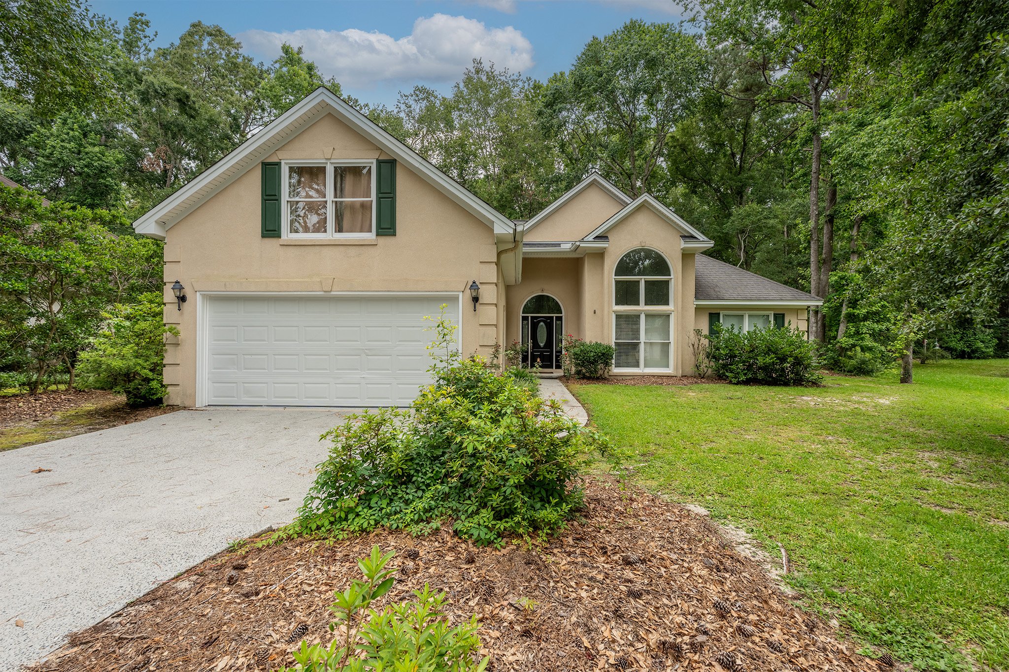 Front view of a beige two-story house with a white garage door, black front door, green shutters, and a large arched window, surrounded by green trees and lawn.
