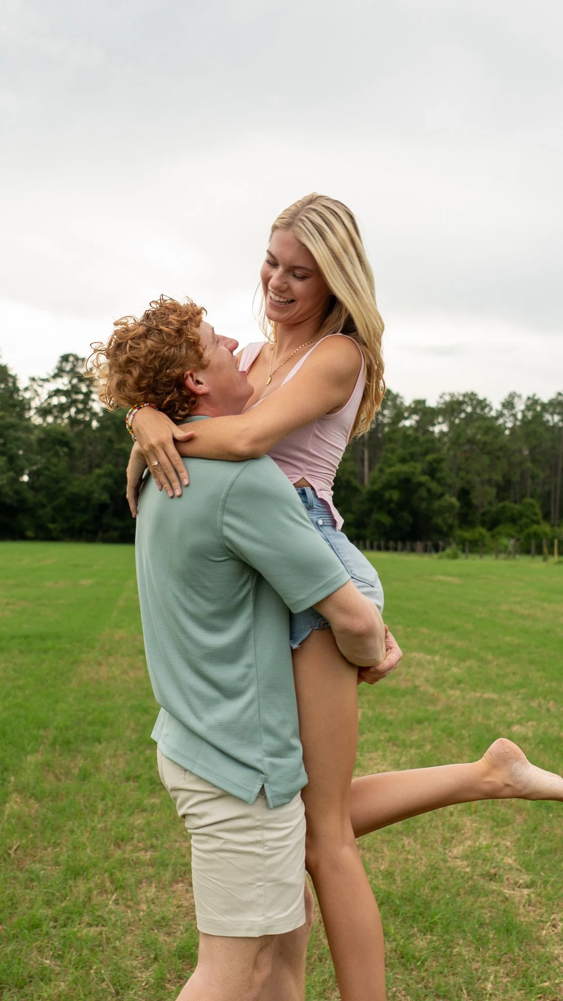 A man lifting a woman in a grassy field with trees in the background, both smiling andembracing.