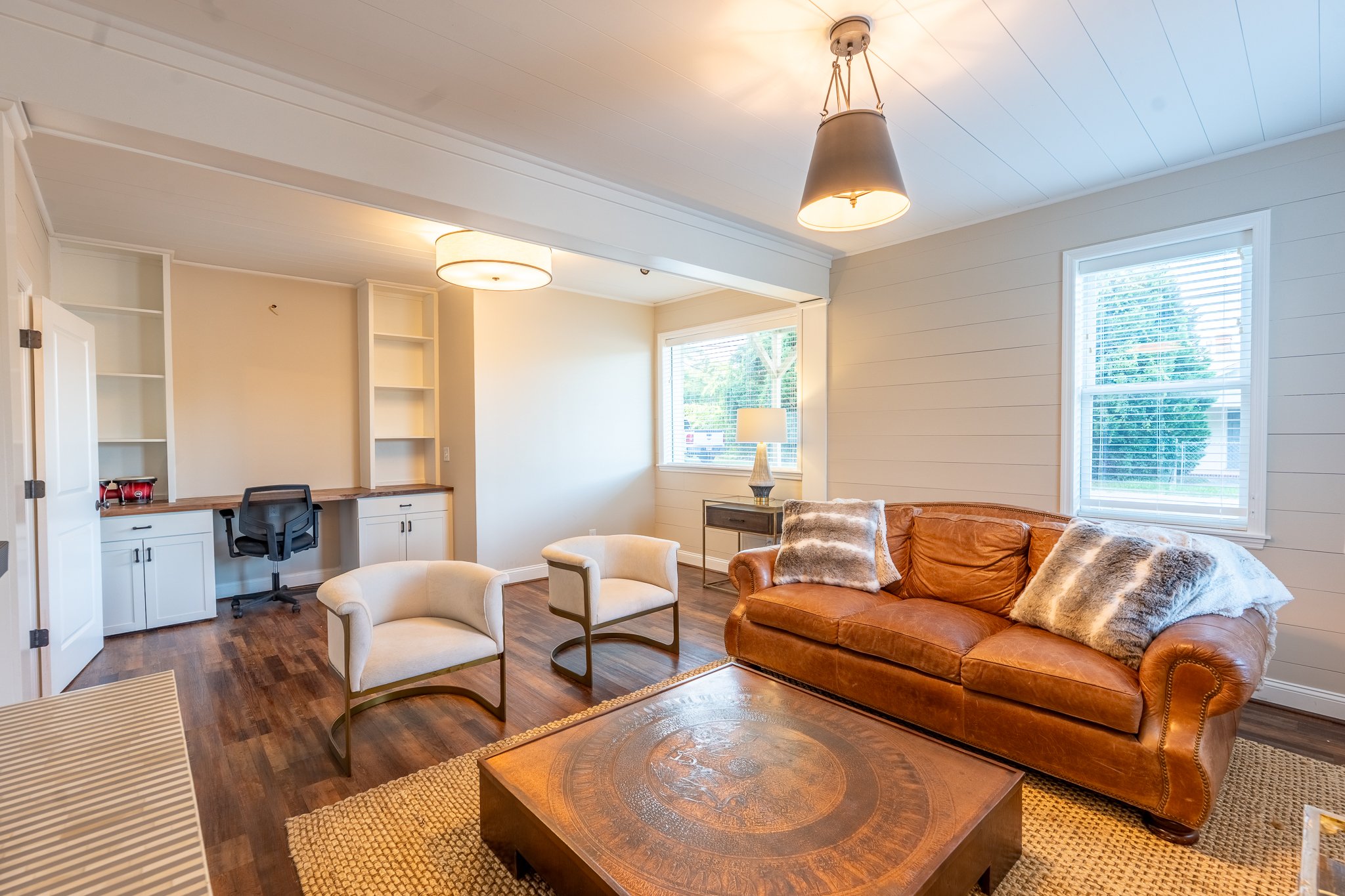 Living room with white walls, wooden floors, a brown leather sofa with fur pillows, two white armchairs, a coffee table, and a window with blinds.