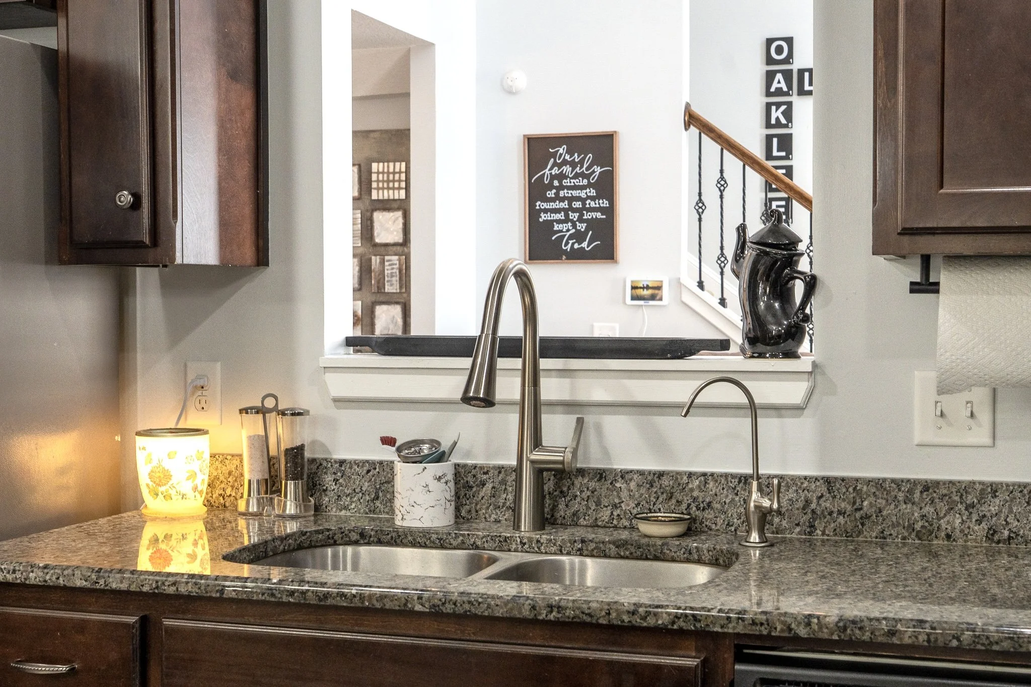 Kitchen sink with granite countertop and dark wood cabinets, view through a pass-through window to a living room with wall decor and staircase.