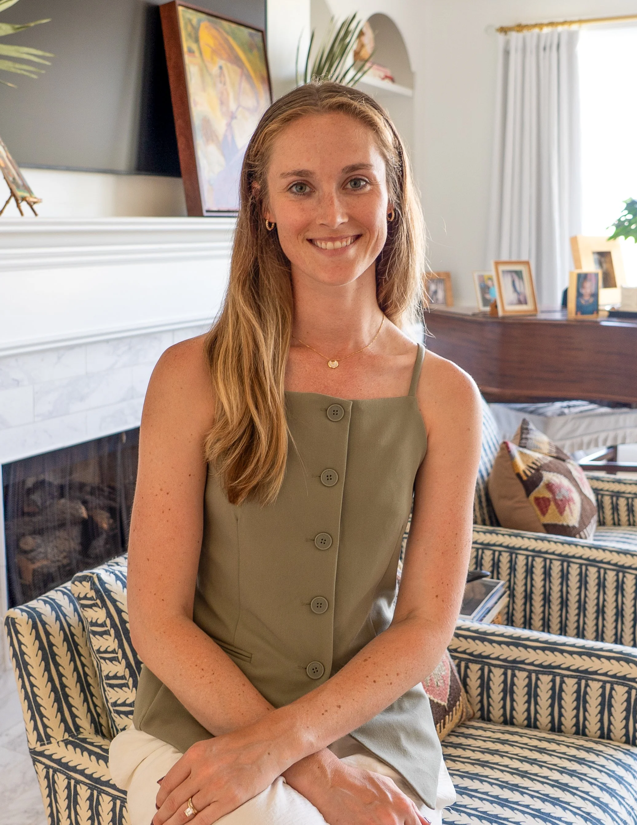 A smiling woman with long, light brown hair sitting on a blue and white patterned couch in a well-lit living room, with framed photographs and artwork in the background.