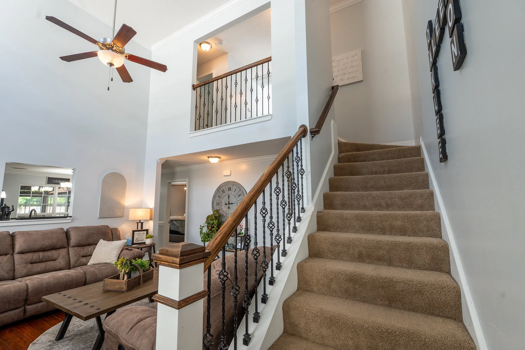 View of living room with staircase leading to upper floor, brown carpeted stairs, wooden handrail, beige walls decorated with black framed words, sofa, clock, and plants.