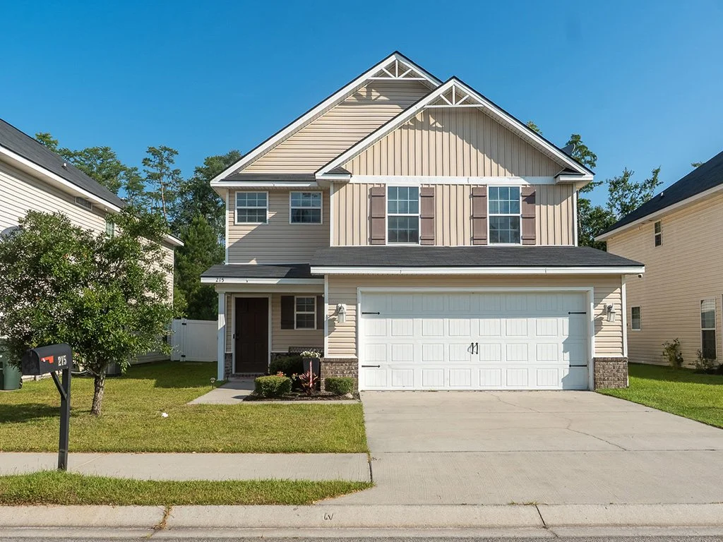 A two-story beige house with brown shutters, a white garage door, and a small front porch, situated on a well-maintained lawn with a tree to the left, under a clear blue sky.