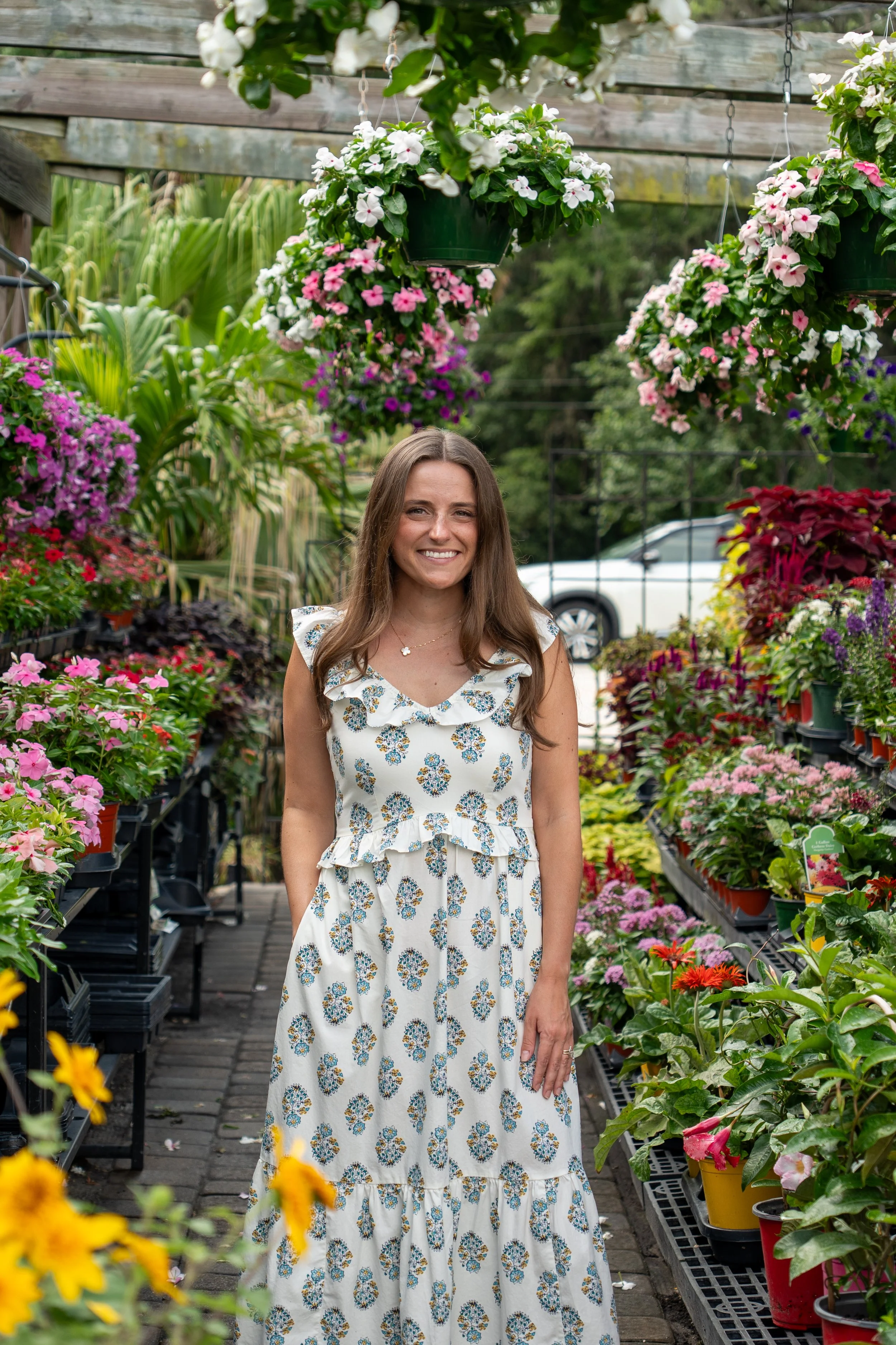 A woman with long brown hair wearing a white floral dress smiling in a garden center among colorful potted flowers on shelves.