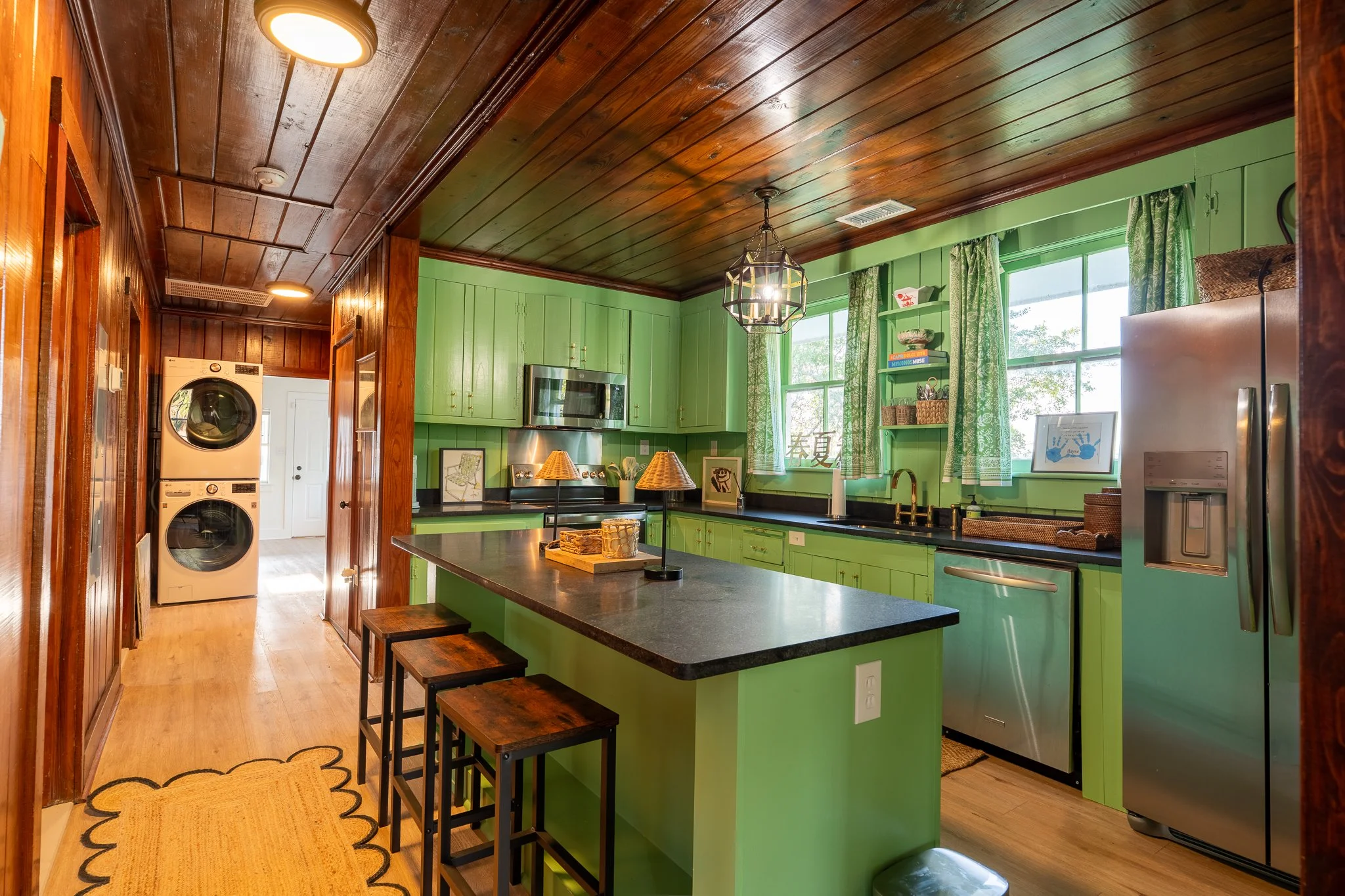 Colorful kitchen with green cabinets, wooden ceiling, black countertops, stainless steel appliances, a kitchen island with seating, and large windows with green curtains.