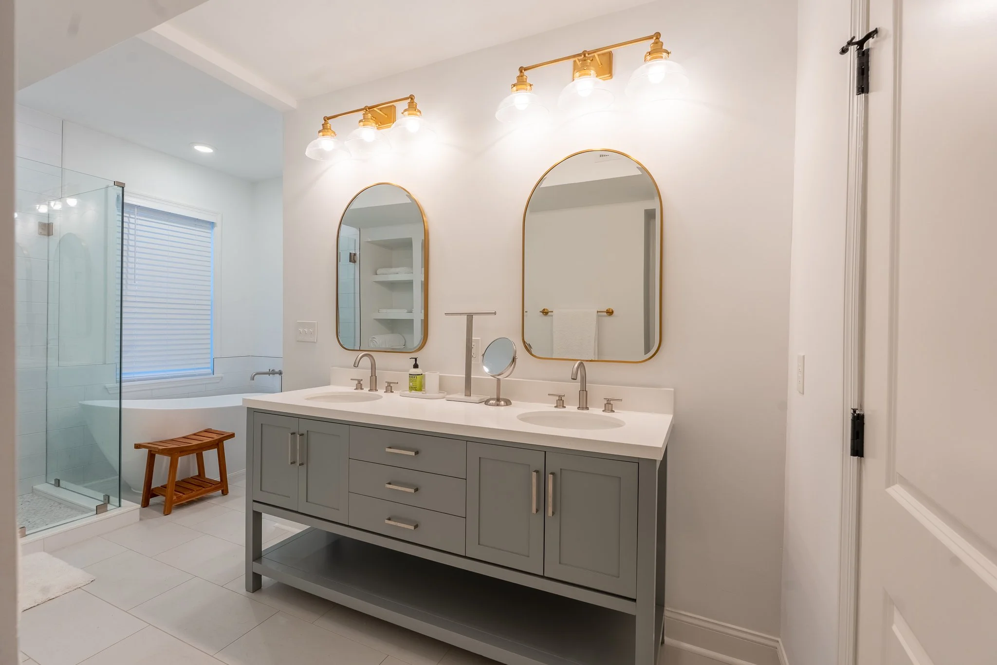 Modern bathroom with double vanity, two mirrors, three-light fixture, and a glass-enclosed shower with a small wooden stool nearby.