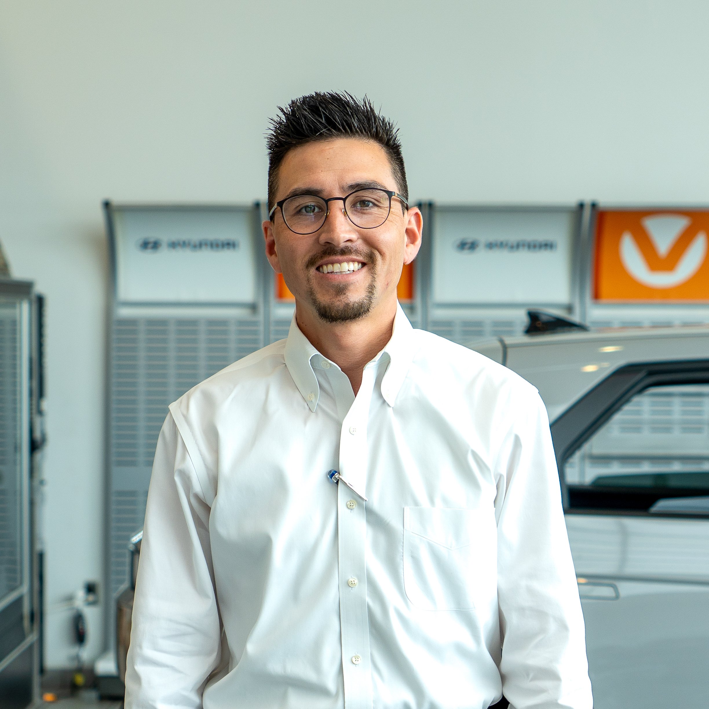 A smiling man with glasses and a goatee is wearing a white button-up shirt and standing inside a car dealership showroom with Hyundai signage in the background.
