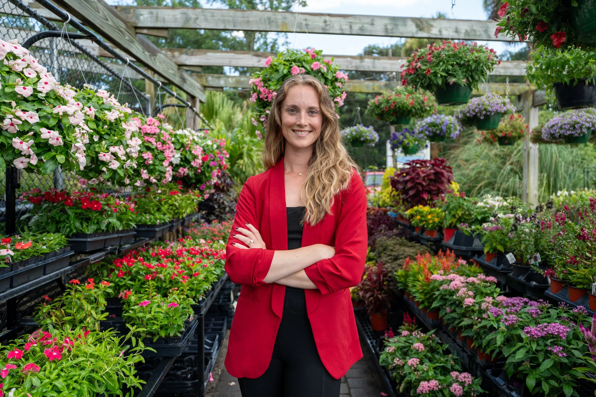 A woman with long wavy blonde hair, wearing a red blazer over a black top, standing with arms crossed in a greenhouse or garden center filled with colorful potted flowers and hanging baskets.