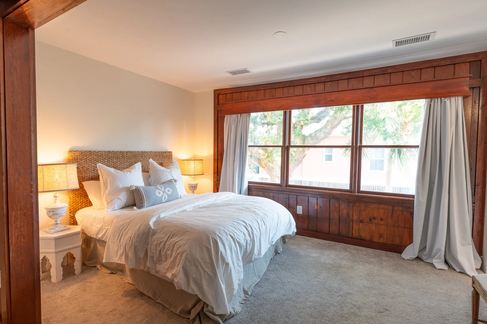 A cozy bedroom featuring a bed with white bedding, decorative pillows, and two bedside lamps. Large window with wooden trim and white curtains overlooks tree branches outside.