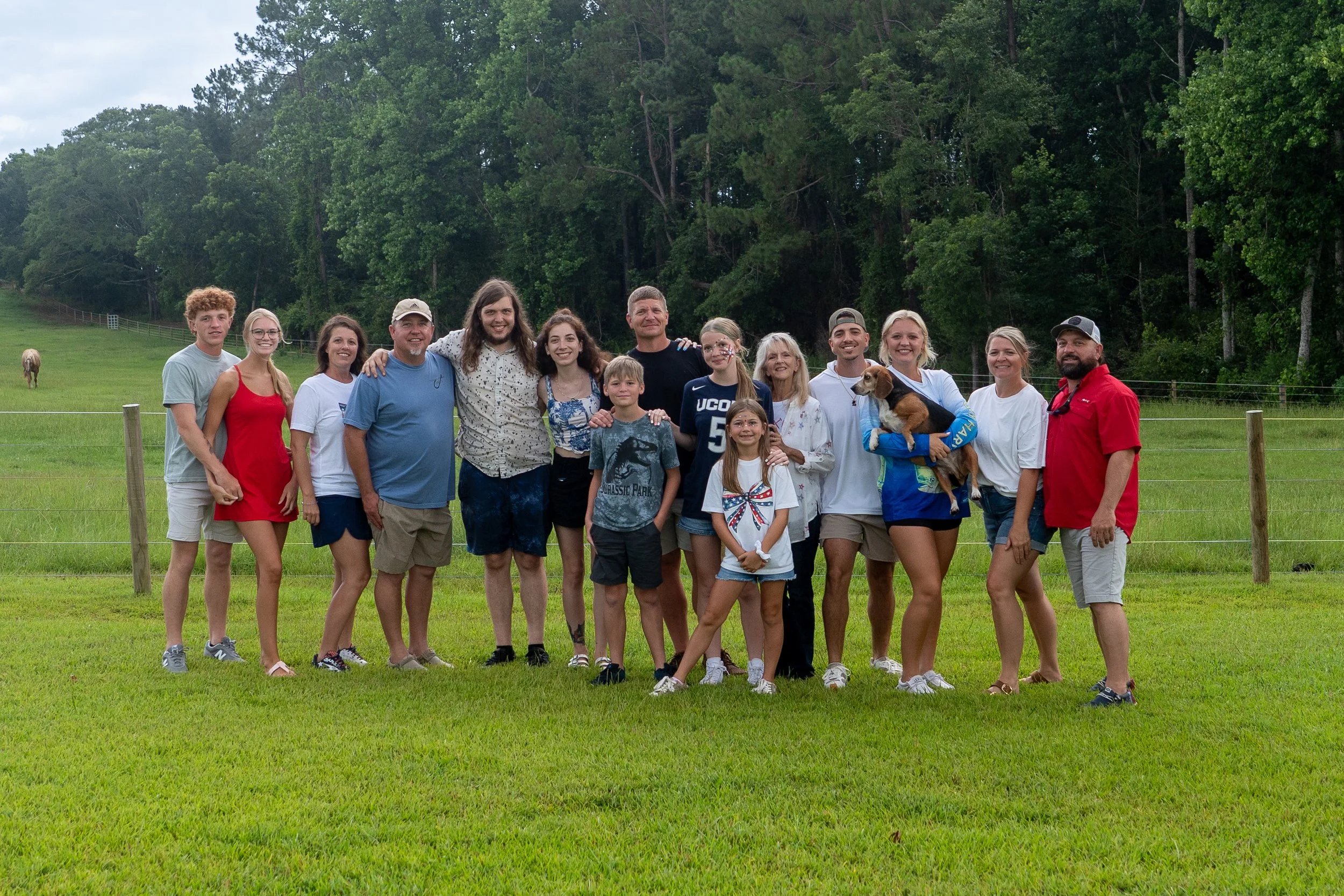 A large group of people standing outdoors in a grassy area with trees in the background. They are smiling and posing for a photo, with some children and a dog included in the group.