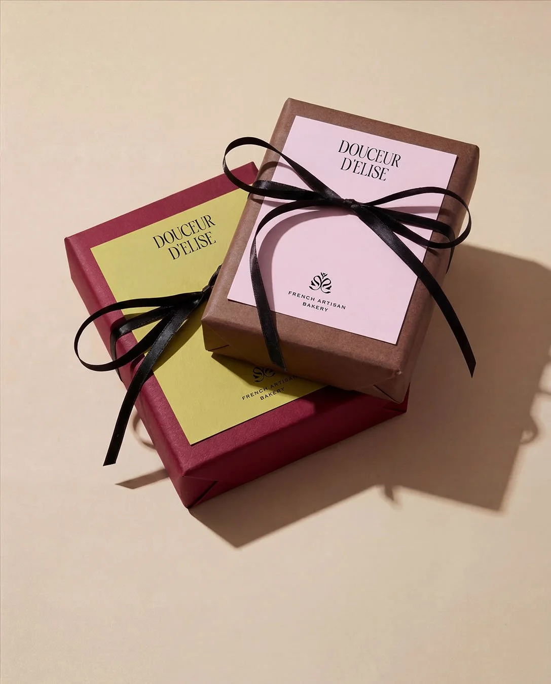 Two gift boxes wrapped in paper, one with a pink label and the other with a yellow label, both labeled 'DOUCEUR D'ELISE' from French Artisan Bakery, tied with black ribbons.
