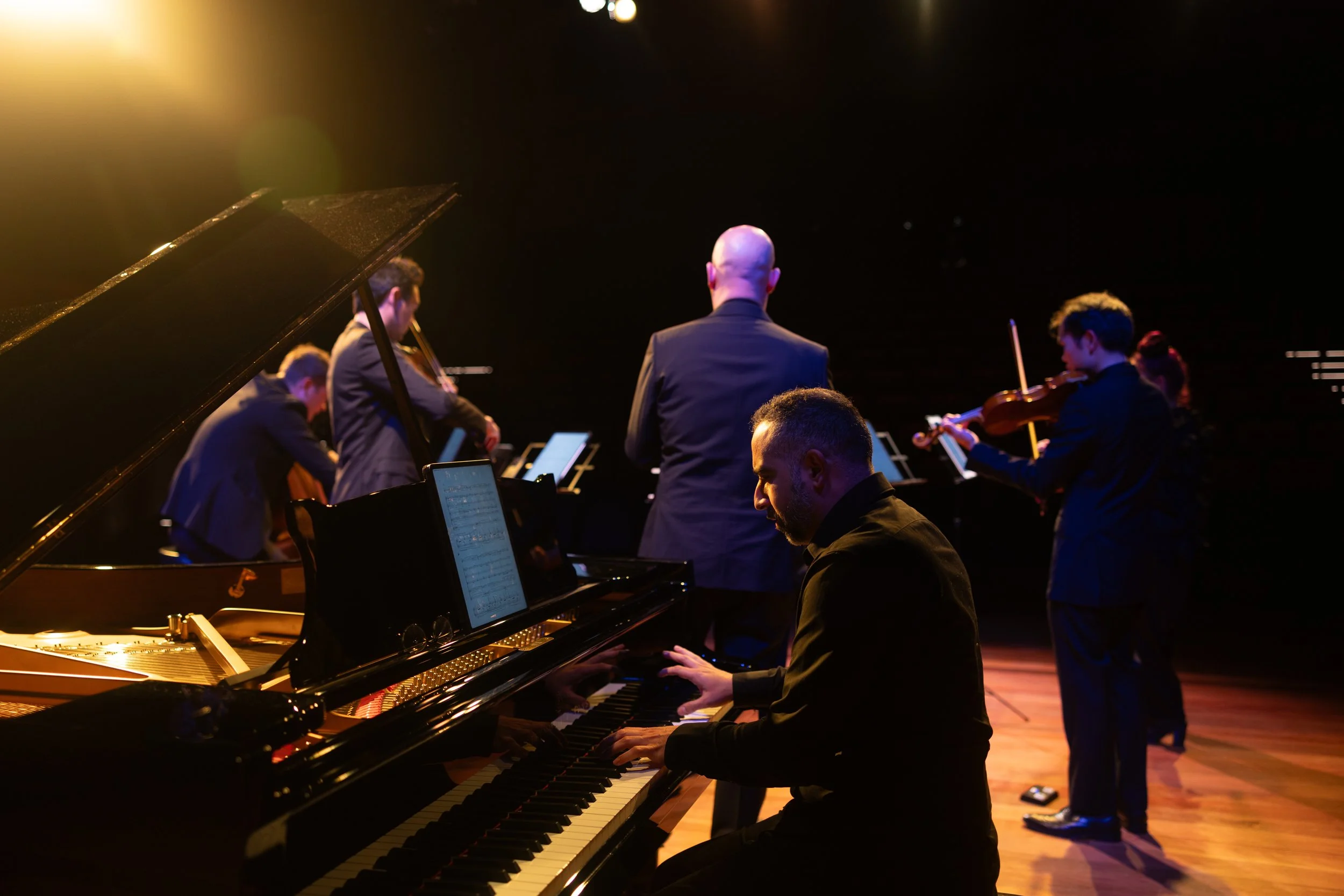 Orchestra performing on stage with pianist in foreground and violinists in background, dim lighting, wooden stage.