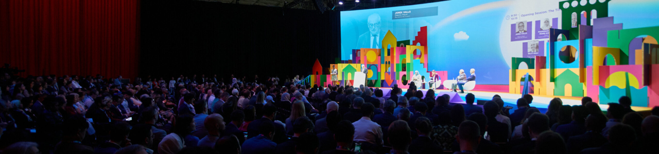 Auditorio lleno durante una conferencia de Smart City, con panelistas en un escenario decorado como una ciudad de formas geométricas y colores brillantes.
