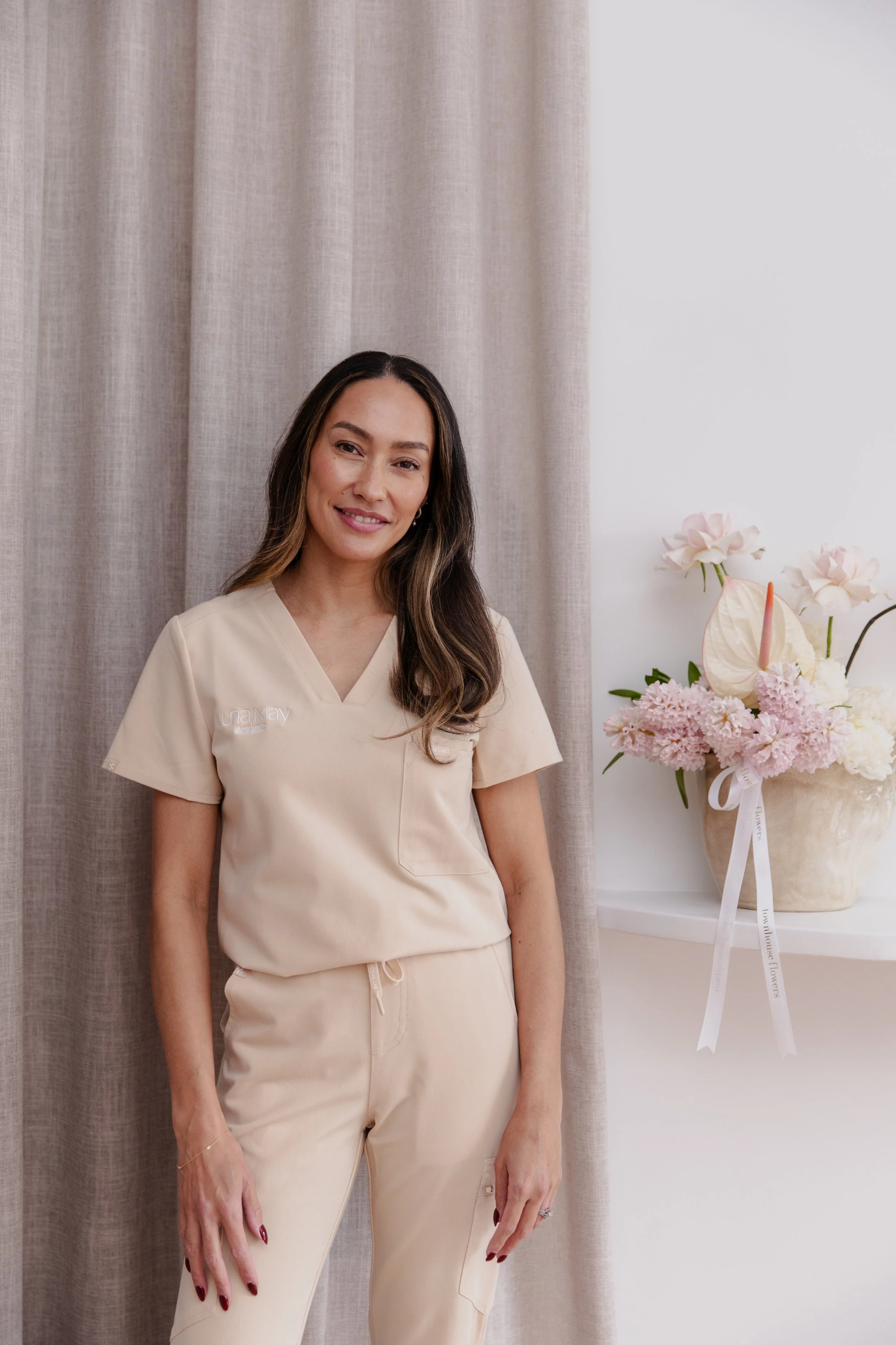 A woman with long, wavy hair and light makeup smiling in a beige medical scrub top and pants standing indoors next to a beige curtain and a floral arrangement in a beige vase with pink and white flowers.