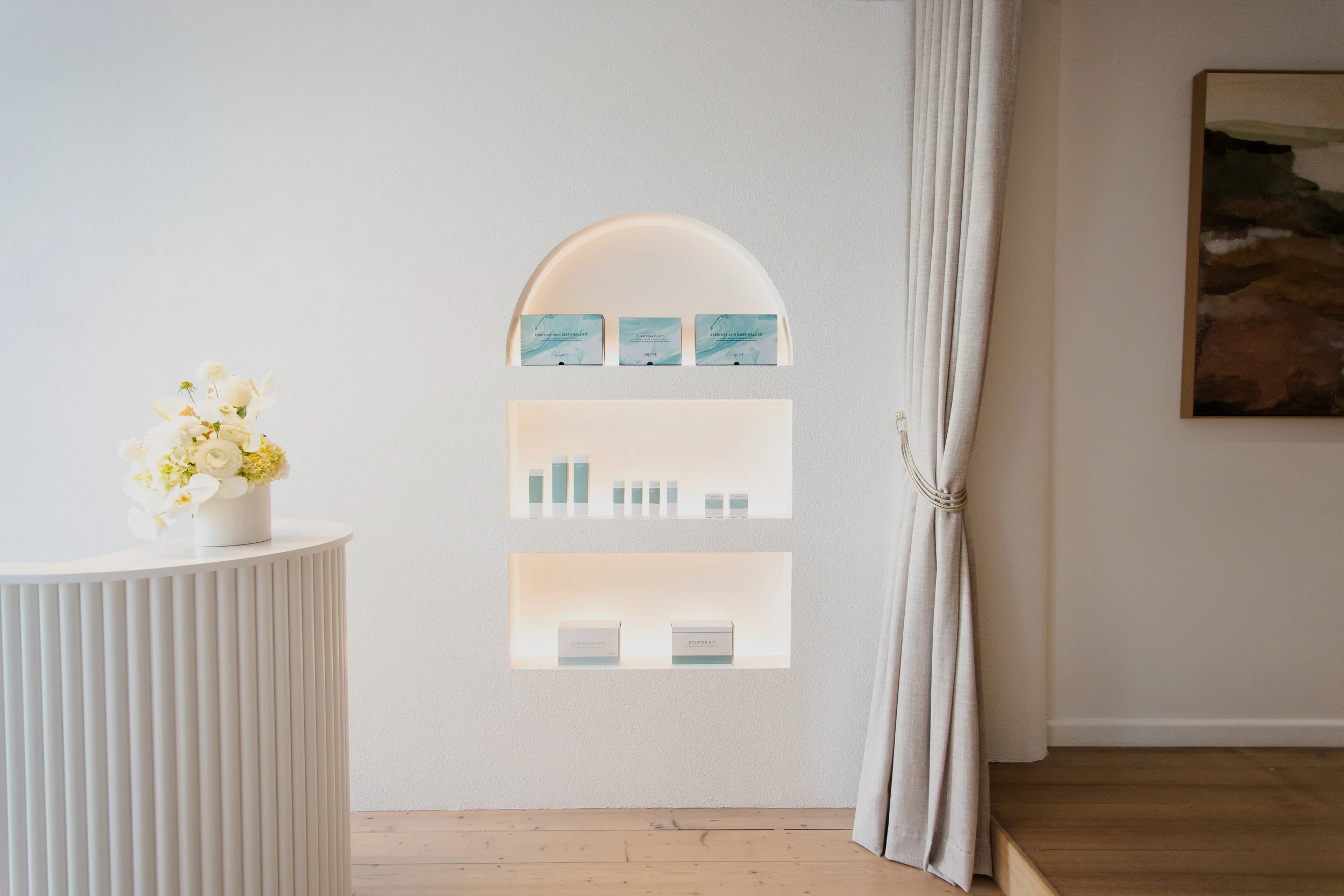 Minimalist interior with built-in wall shelves illuminated with warm light, displaying skincare products, with a white flower arrangement on a white curved counter, beige curtains tied back on the right, and a landscape painting on the wall.