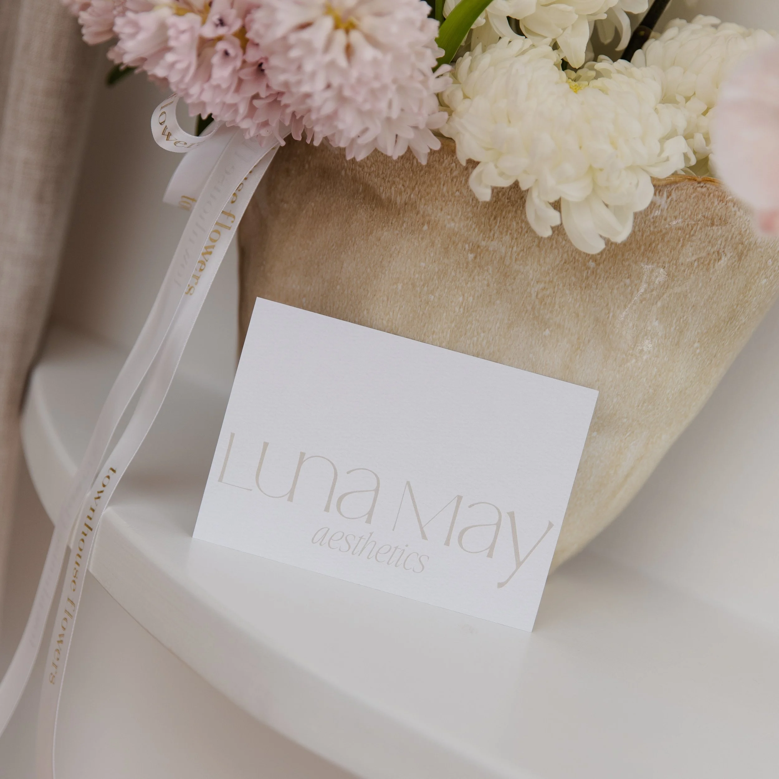 A business card for Luna May Aesthetics placed on a white surface near a beige flowerpot with pink and white flowers.