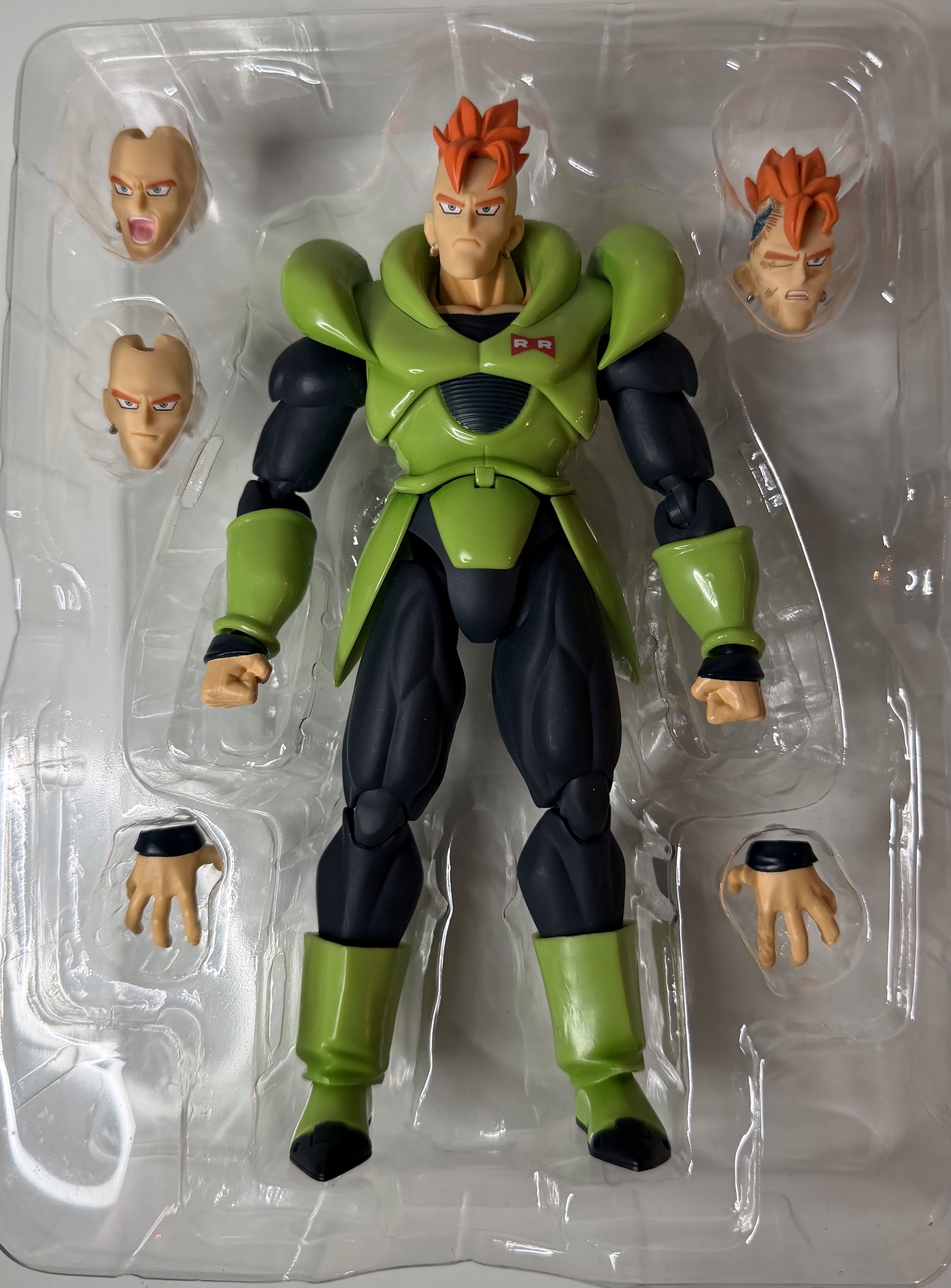 Action figure of a muscular male character with orange hair, wearing green and black armor, with three extra heads and two hands in a plastic packaging.