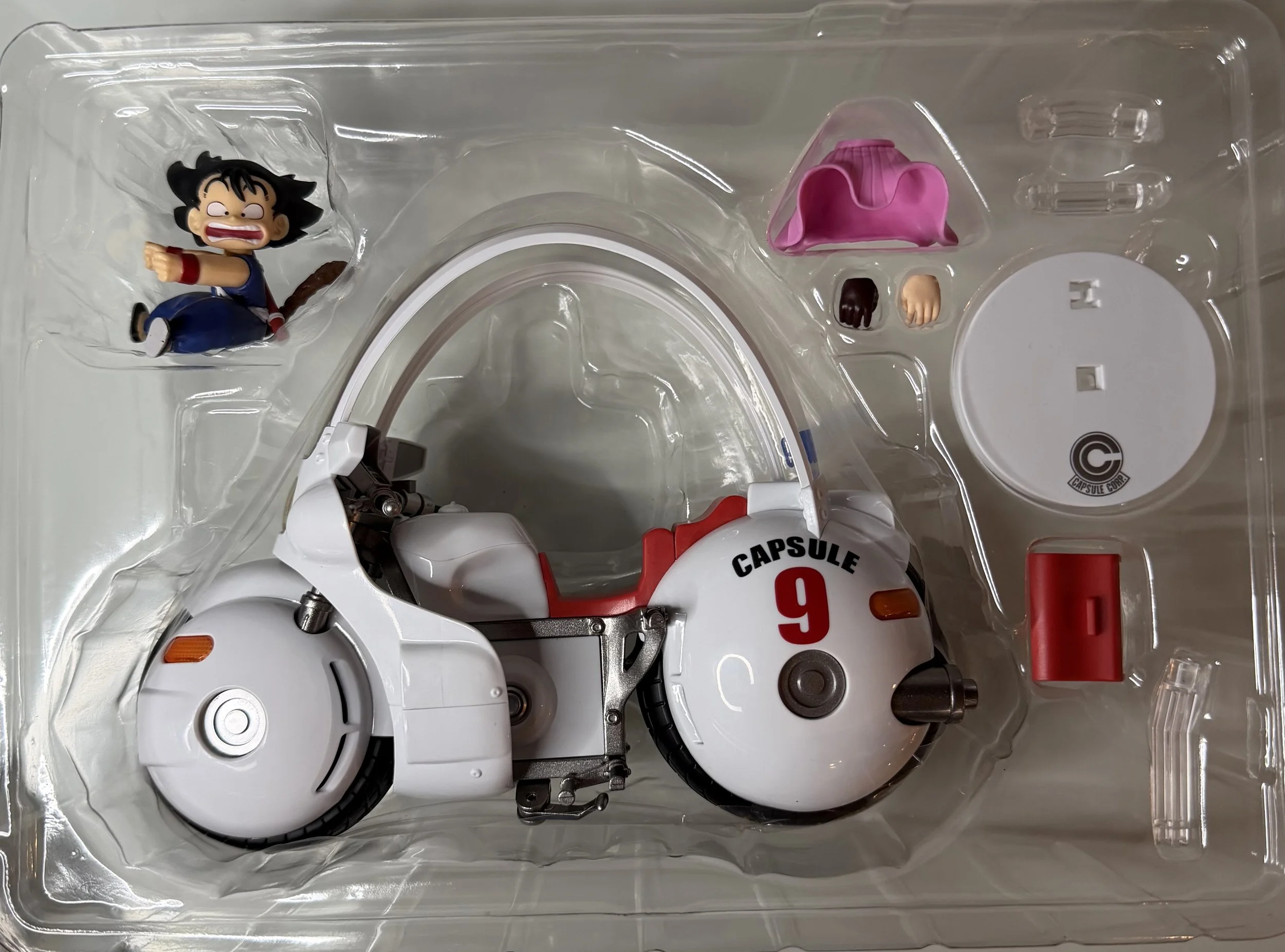A plastic boxed set of a small, white mechanical device with a red and black seat, labeled 'CAPSULE' and '9'. Includes small action figures and accessories like a pink skirt, hands, and other parts.