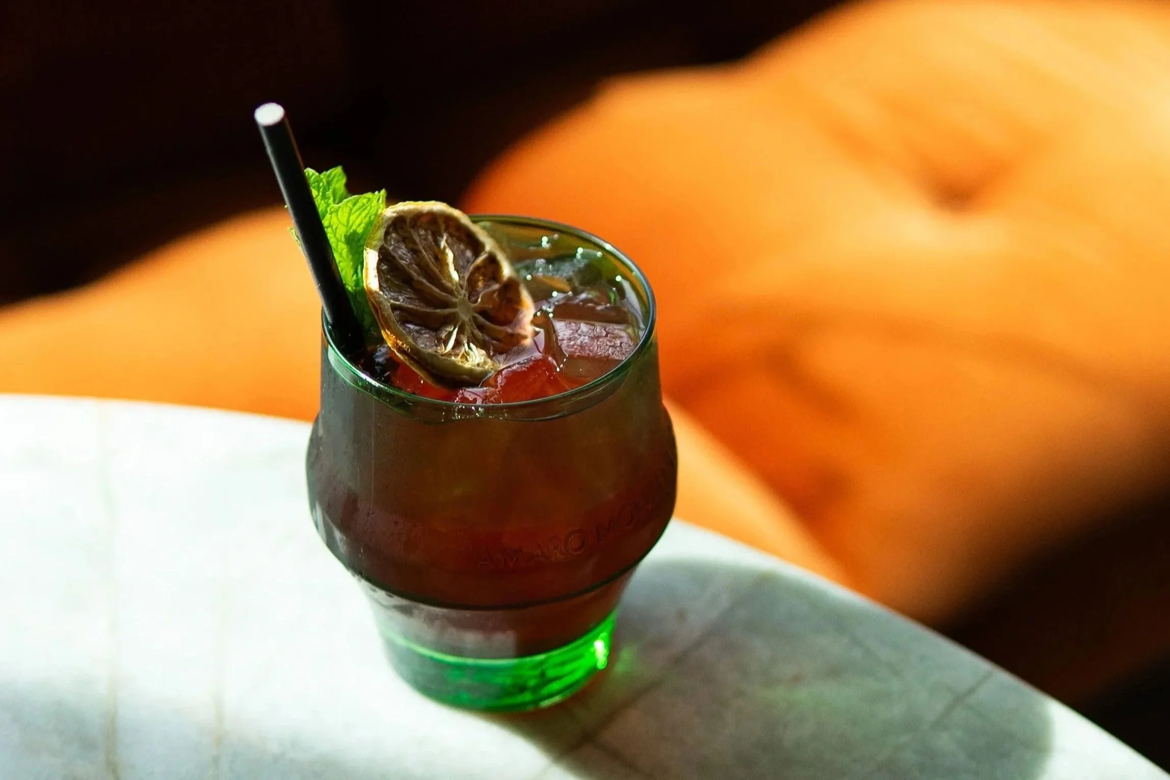 A glass of dark red beverage with a slice of dried lemon and a sprig of mint, with a black straw, on a table with a blurred orange background.