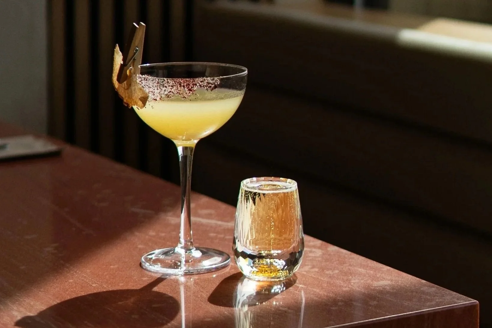 A cocktail glass with a yellow drink garnished with a lemon peel and red sugar rim, alongside a short crystal glass with a golden beverage, on a wooden table with a dark background.