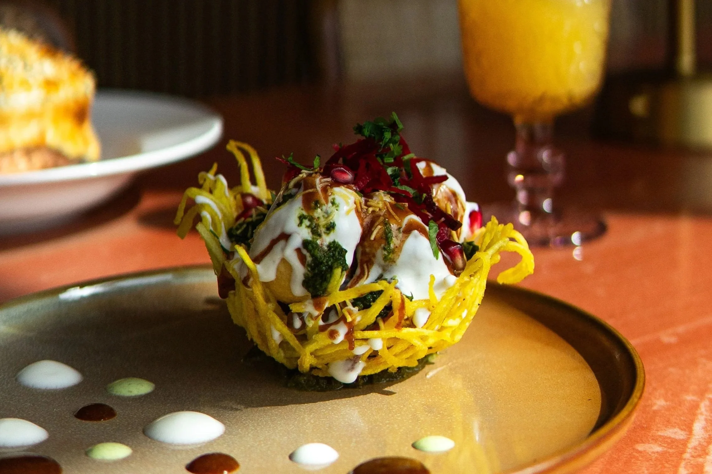 A close-up of a gourmet appetizer featuring a crispy edible cup filled with cream, garnished with pomegranate seeds, chopped herbs, and a drizzle of sauce, served on a black plate with a colorful背景.