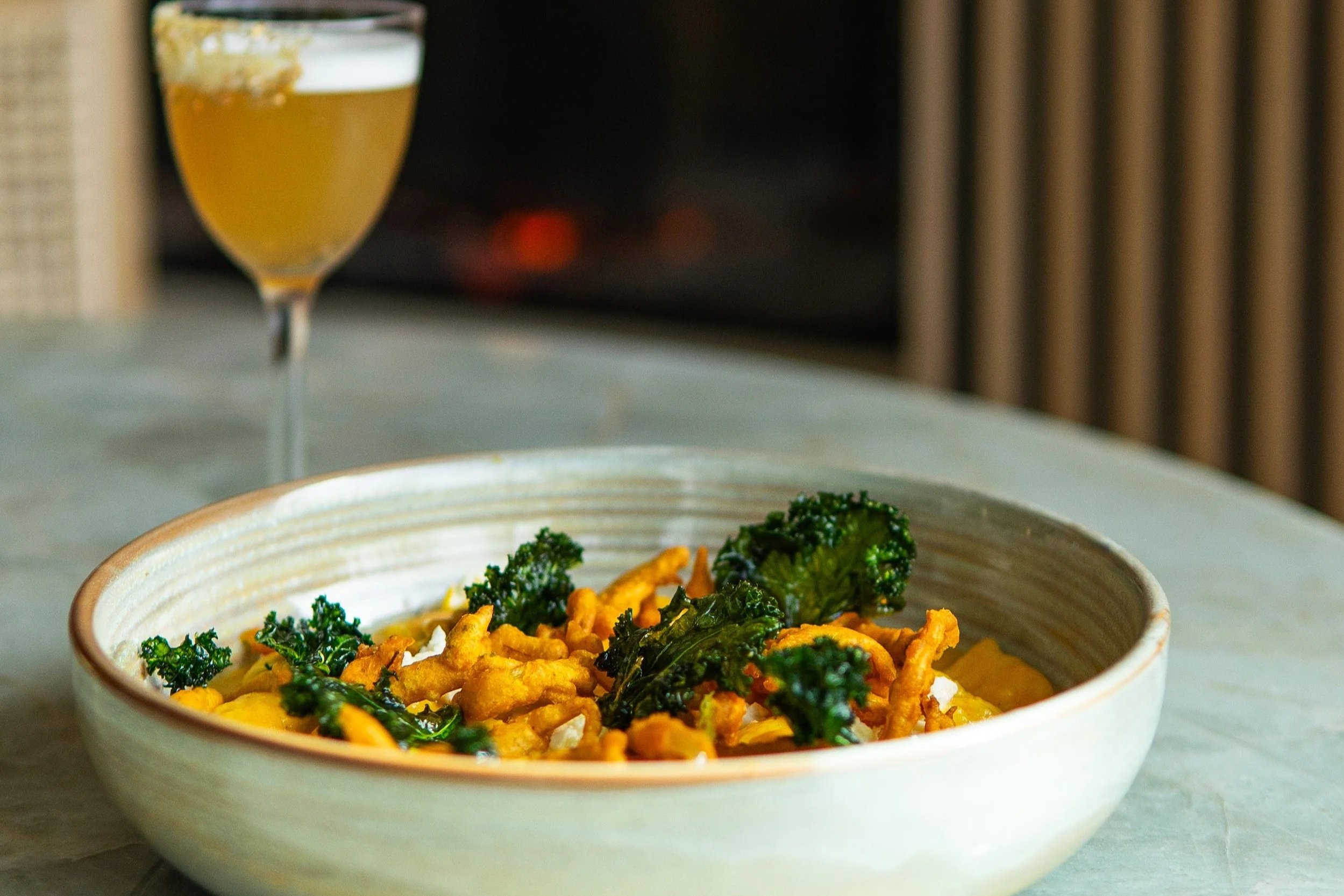 A bowl of fried chicken garnished with kale, placed on a light-colored surface with a blurred background and a glass of beer in the background.