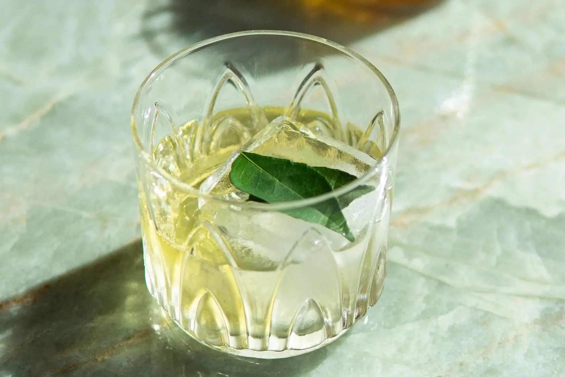 Clear glass with decorative patterns containing a beverage with ice and a green leaf, set on a textured surface.