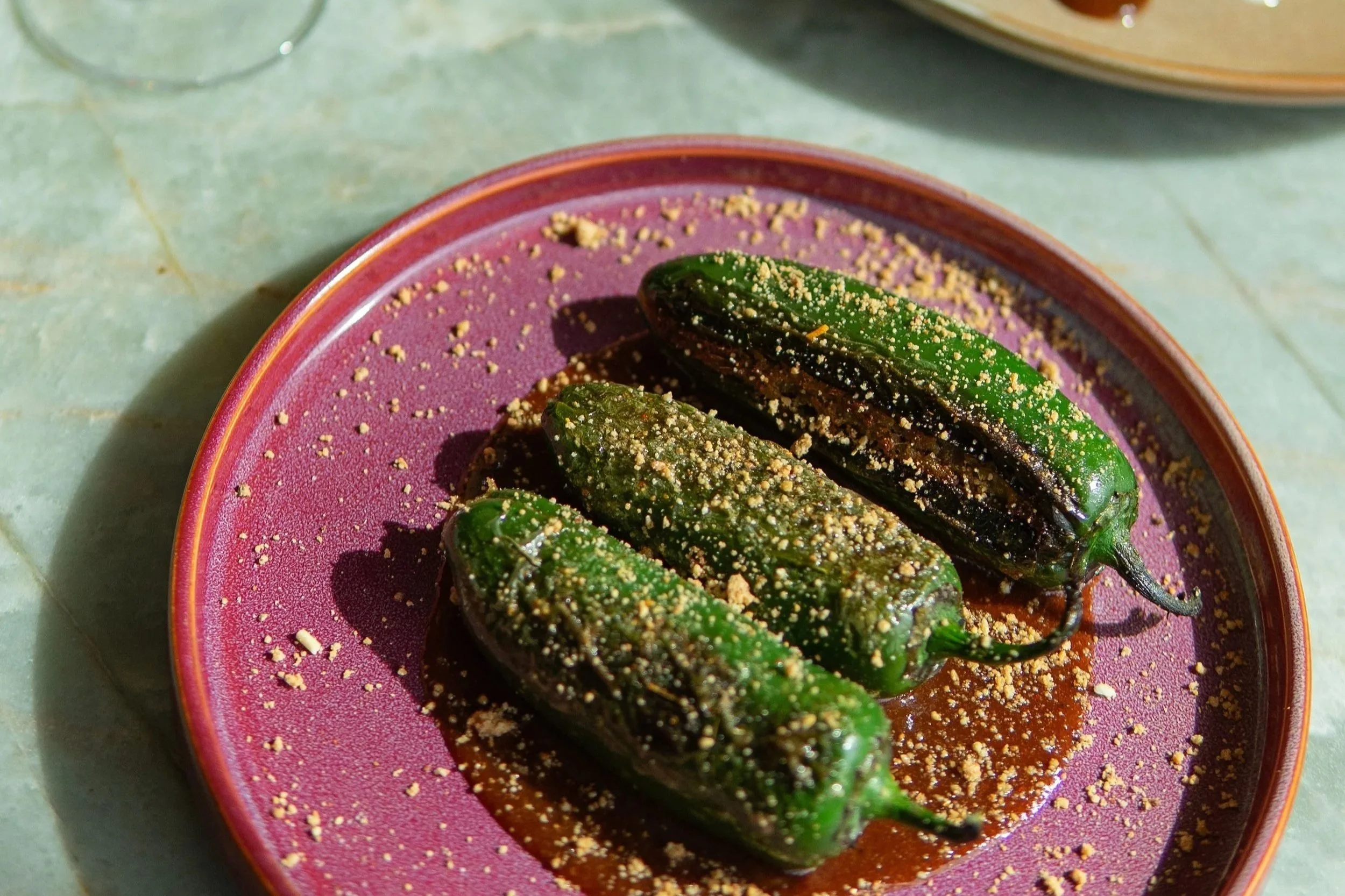 Roasted green chilies sprinkled with crushed nuts on a pink plate.