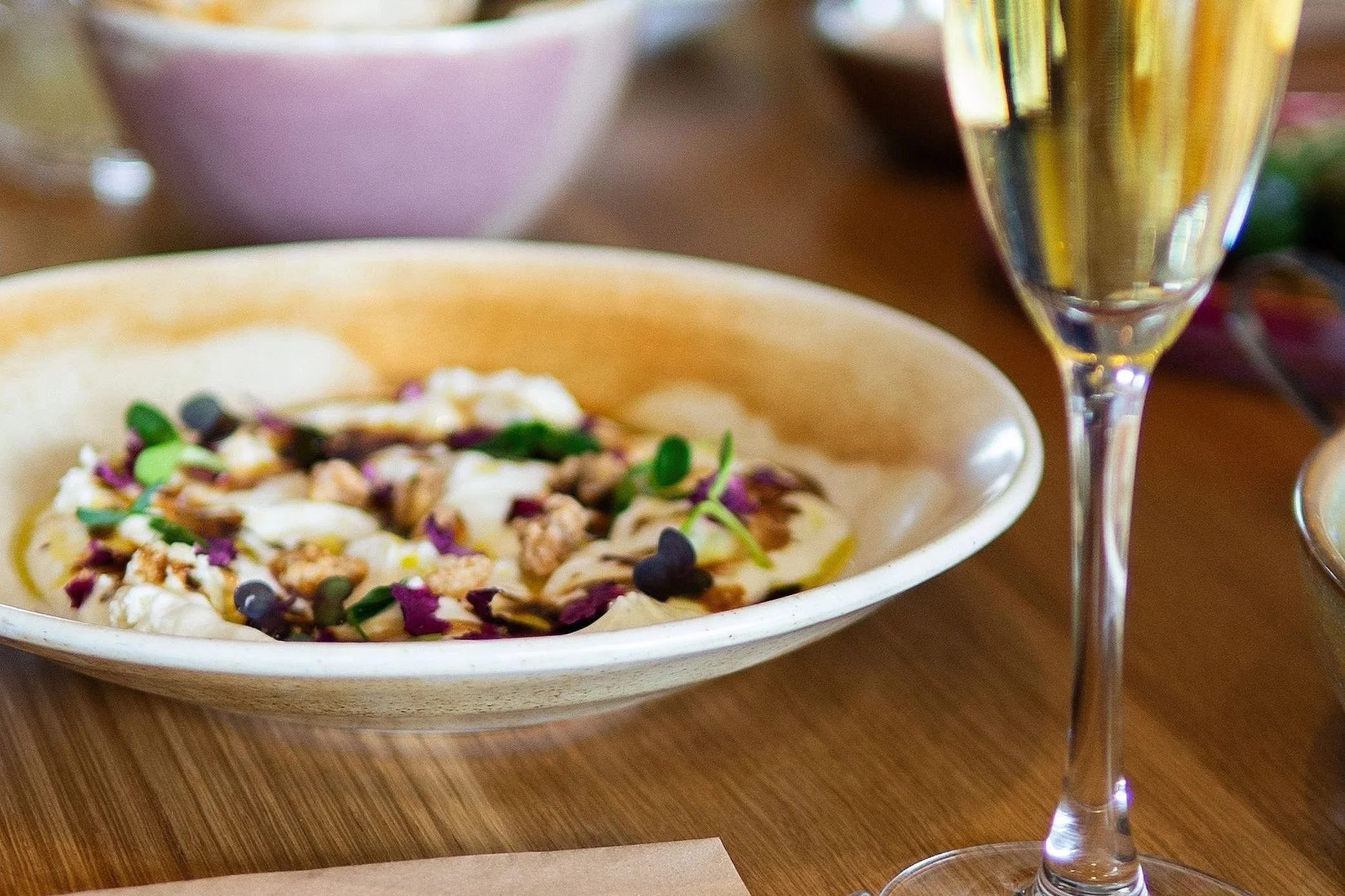 Close-up of a salad with chopped vegetables and microgreens in a white bowl, accompanied by a glass of white wine on a wooden table.