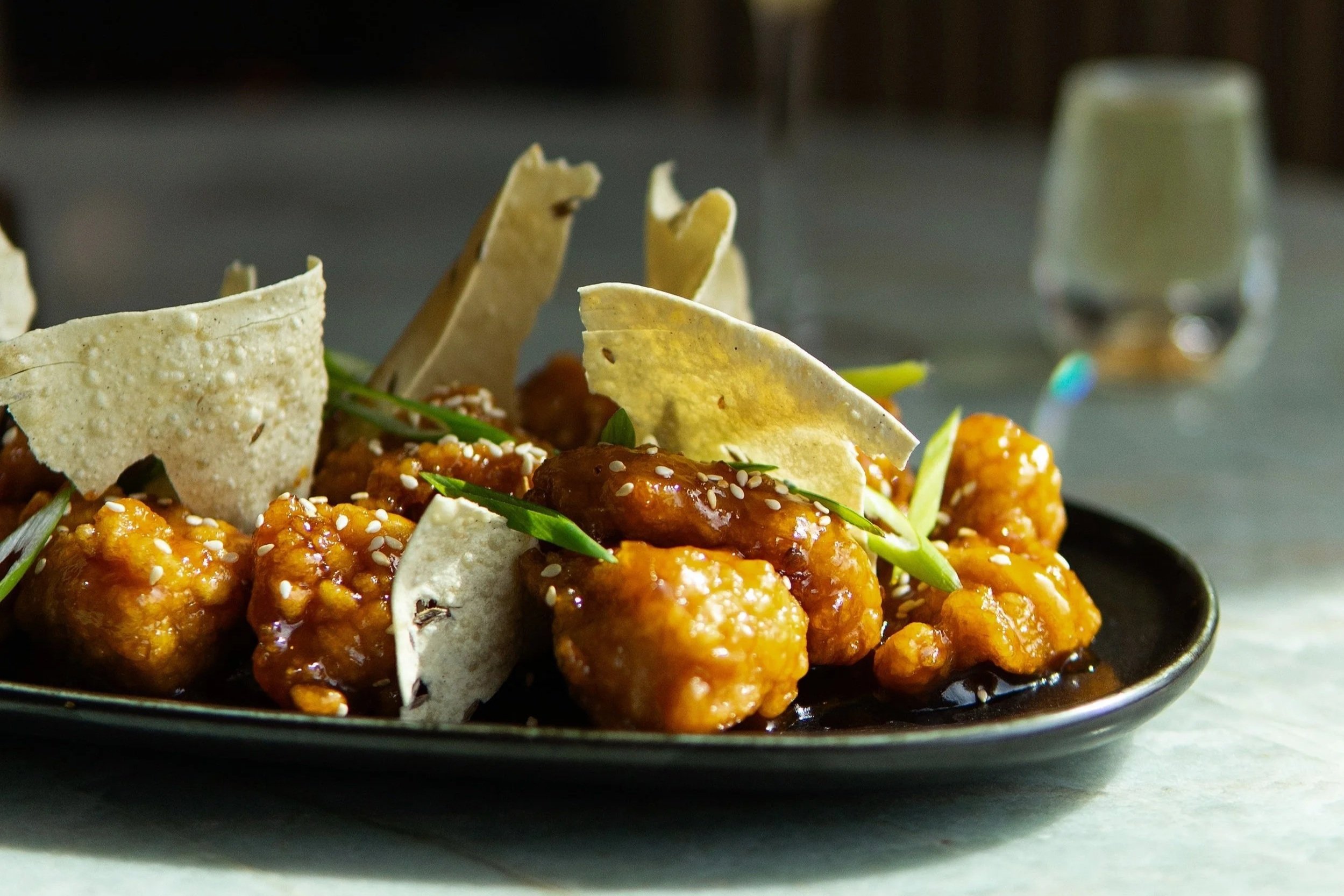 Close-up of spicy glazed chicken bites garnished with sesame seeds, green onions, lettuce, and tortilla chips on a black plate