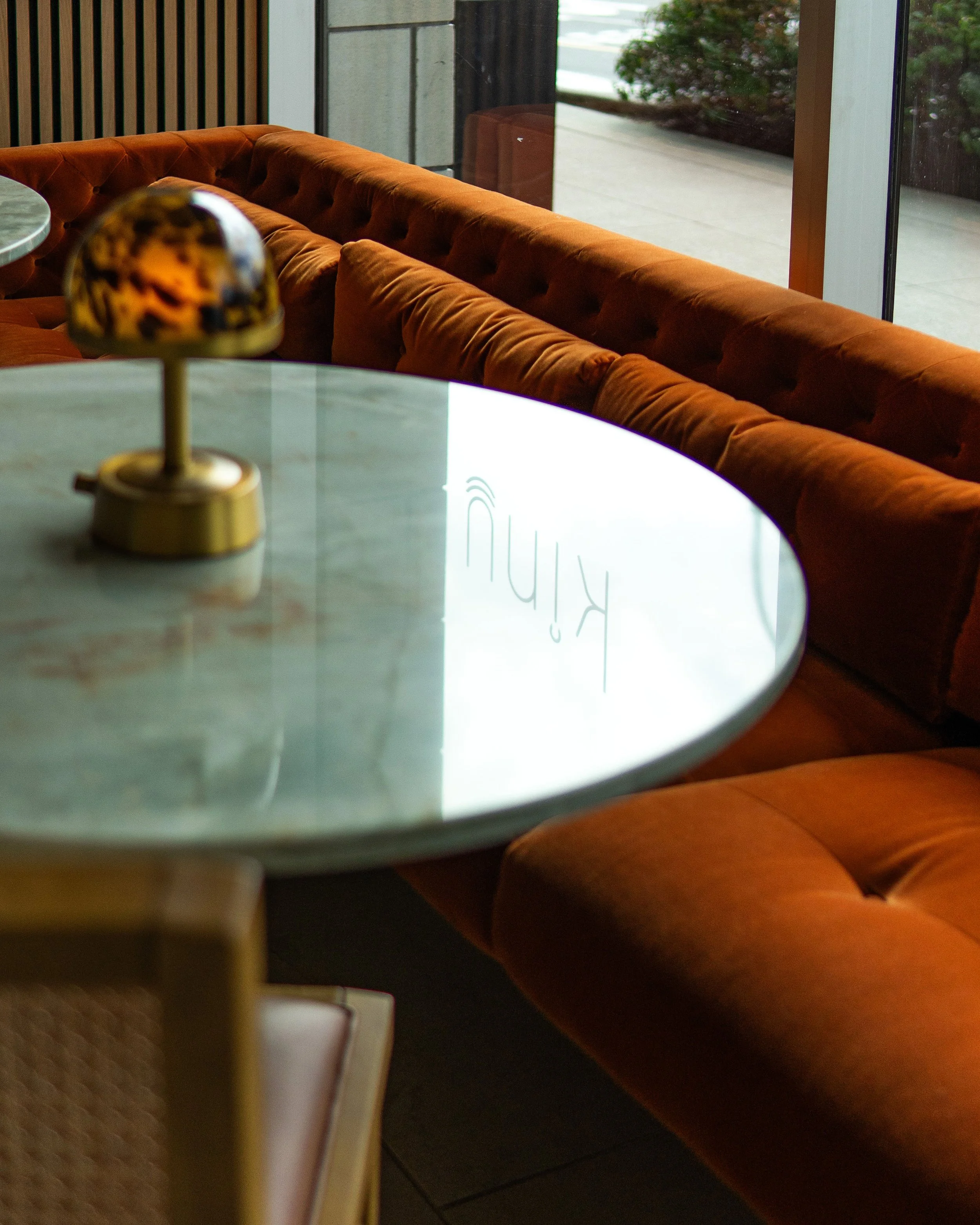 Inside a cafe or restaurant, there are orange tufted upholstered couches placed against a window, with a marble-top table in the foreground, and a decorative lamp with a marble base and colorful shade on the table. Outdoors through the window, greenery and pavement are visible.