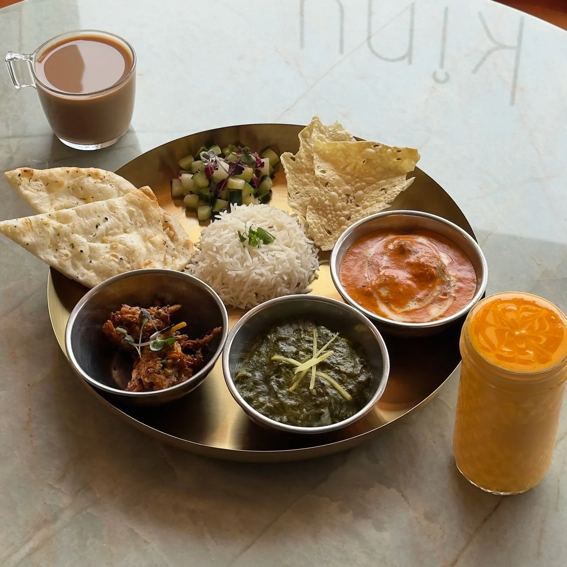 A person holds a plate with a colorful dish, surrounded by other plates of various Indian foods, including naan bread, oysters, rice, and a bowl of curry, on a wooden table.