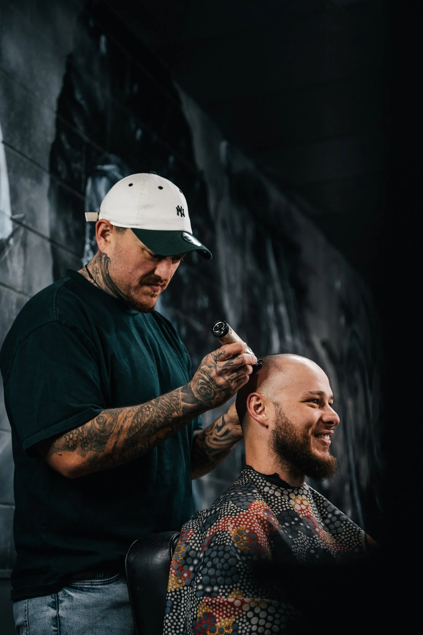 A tattooed man in a baseball cap gives a man with a beard a haircut in a barbershop.