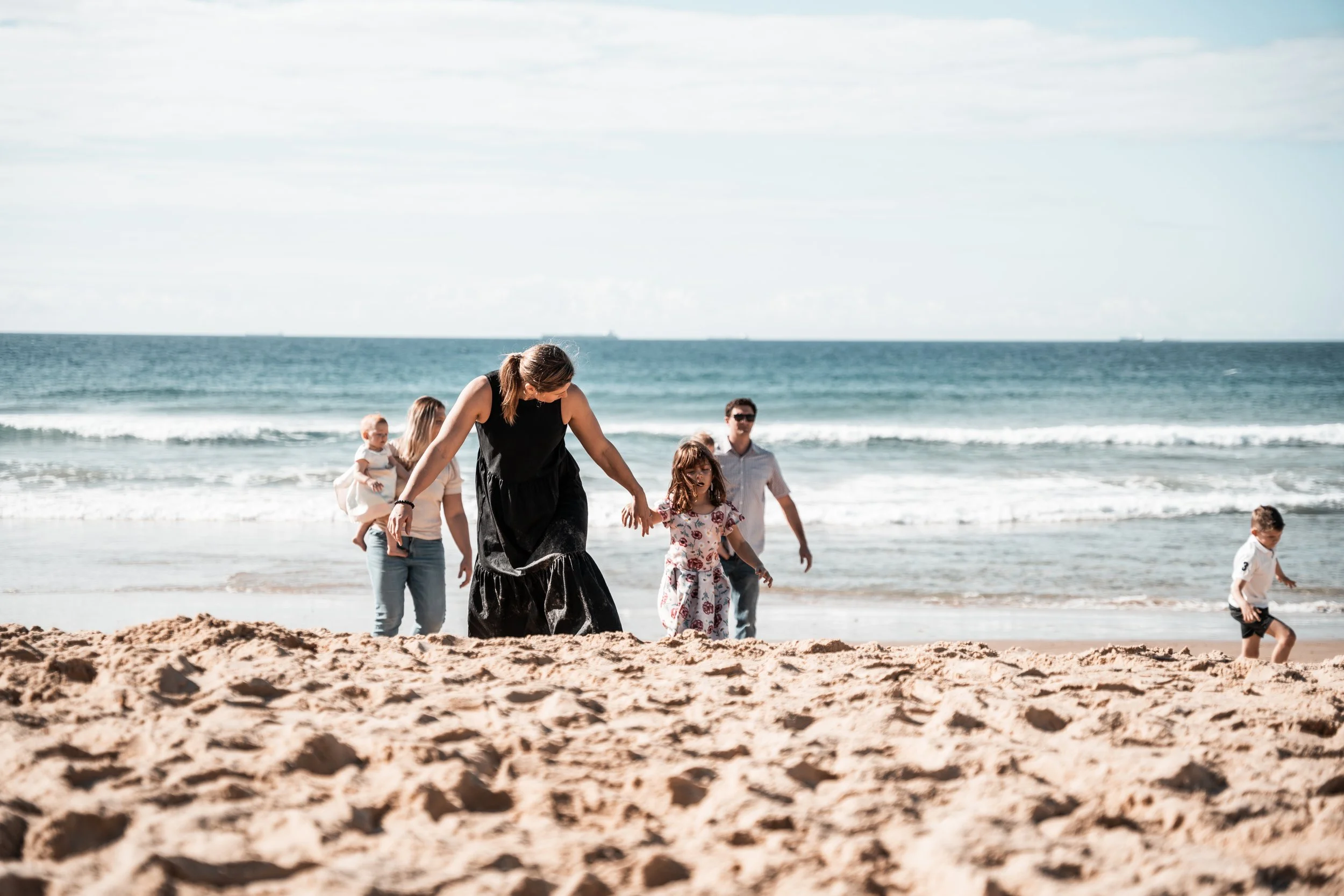 A family of six enjoying a day at the beach, with sand in the foreground and ocean waves in the background. The adults and children are walking and playing near the shoreline.