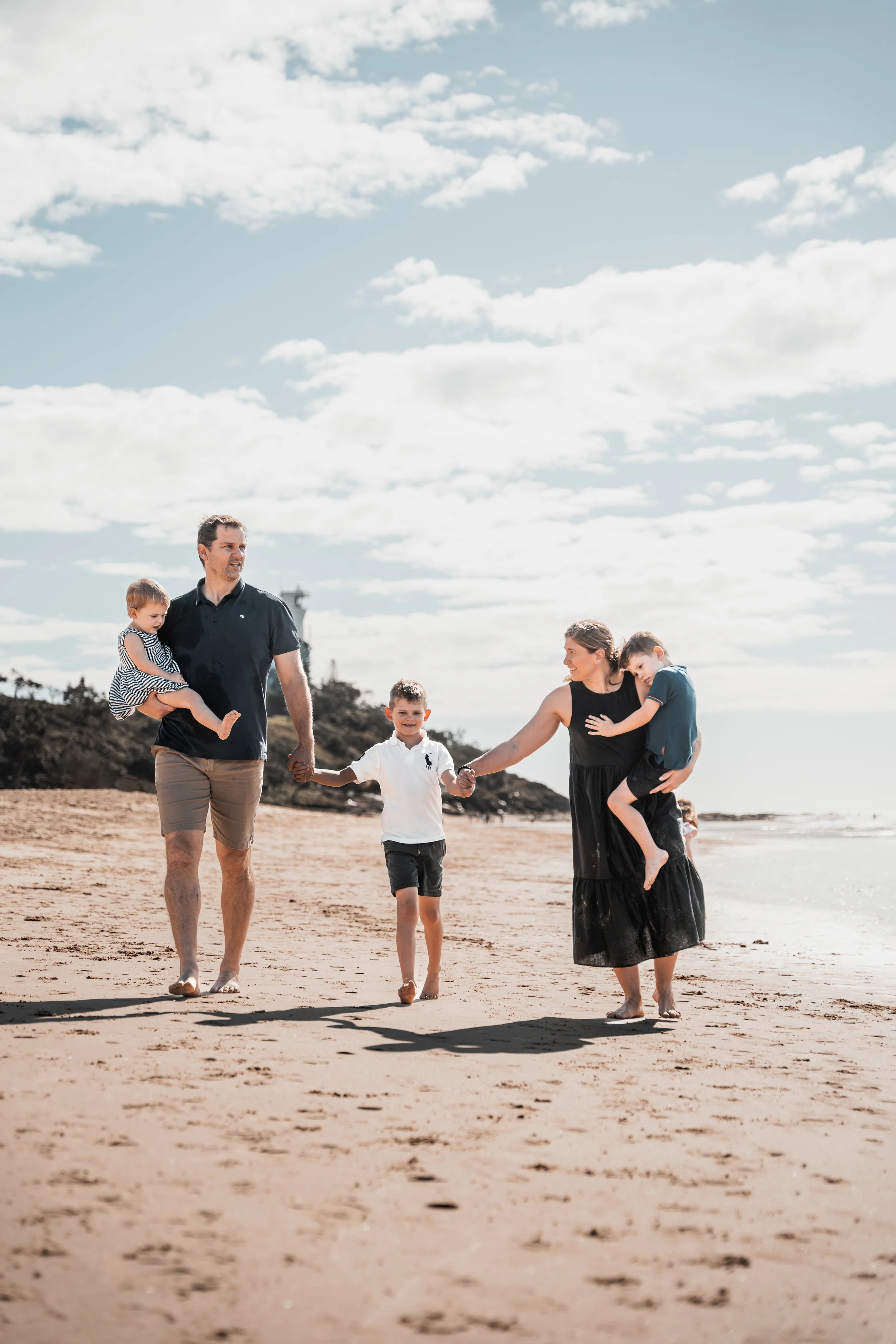 A family of four walking on a sandy beach, holding hands, with a lighthouse in the background and a partly cloudy sky.