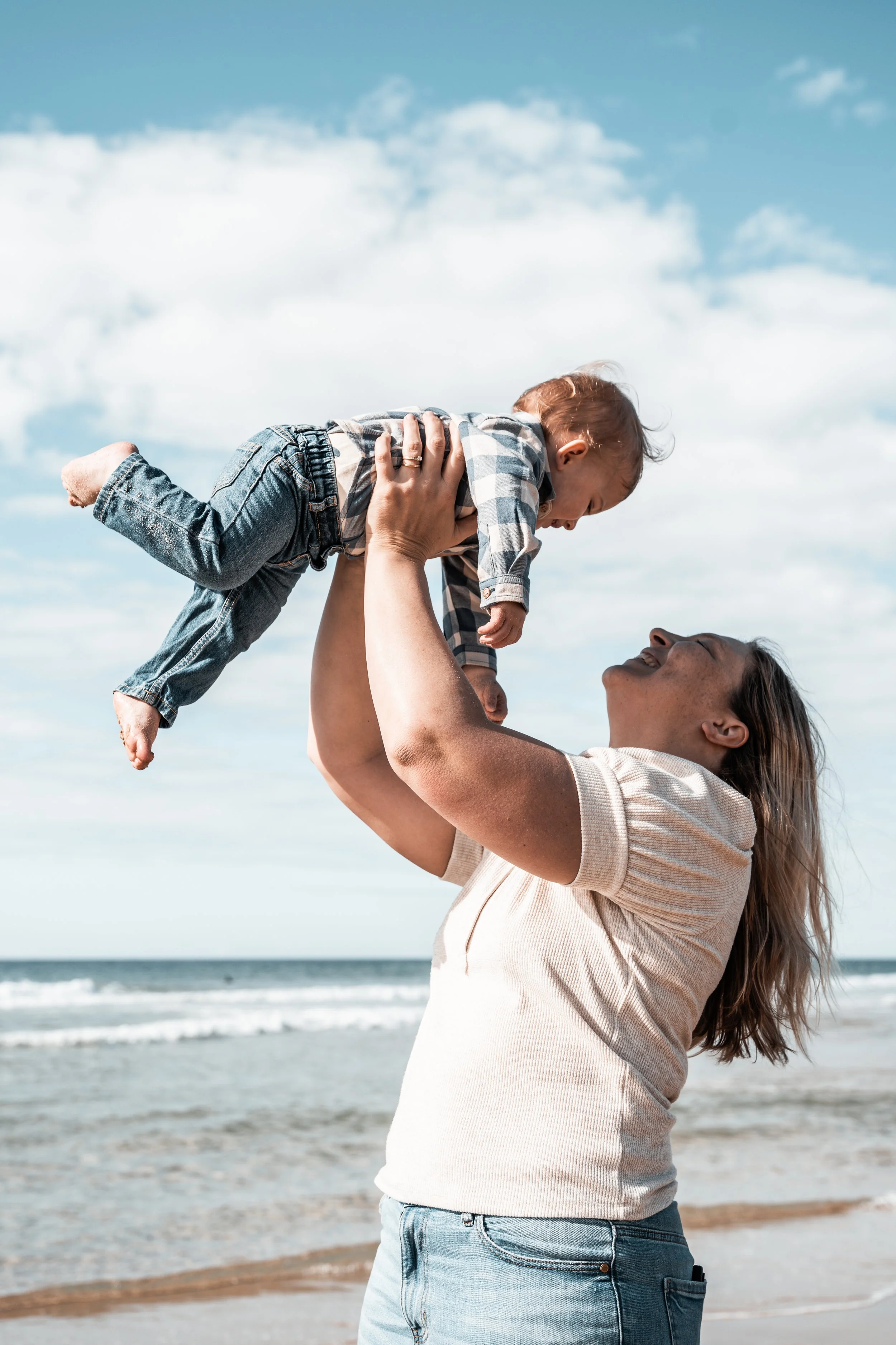 A woman at the beach lifting a young child into the air with a blue sky and ocean in the background.