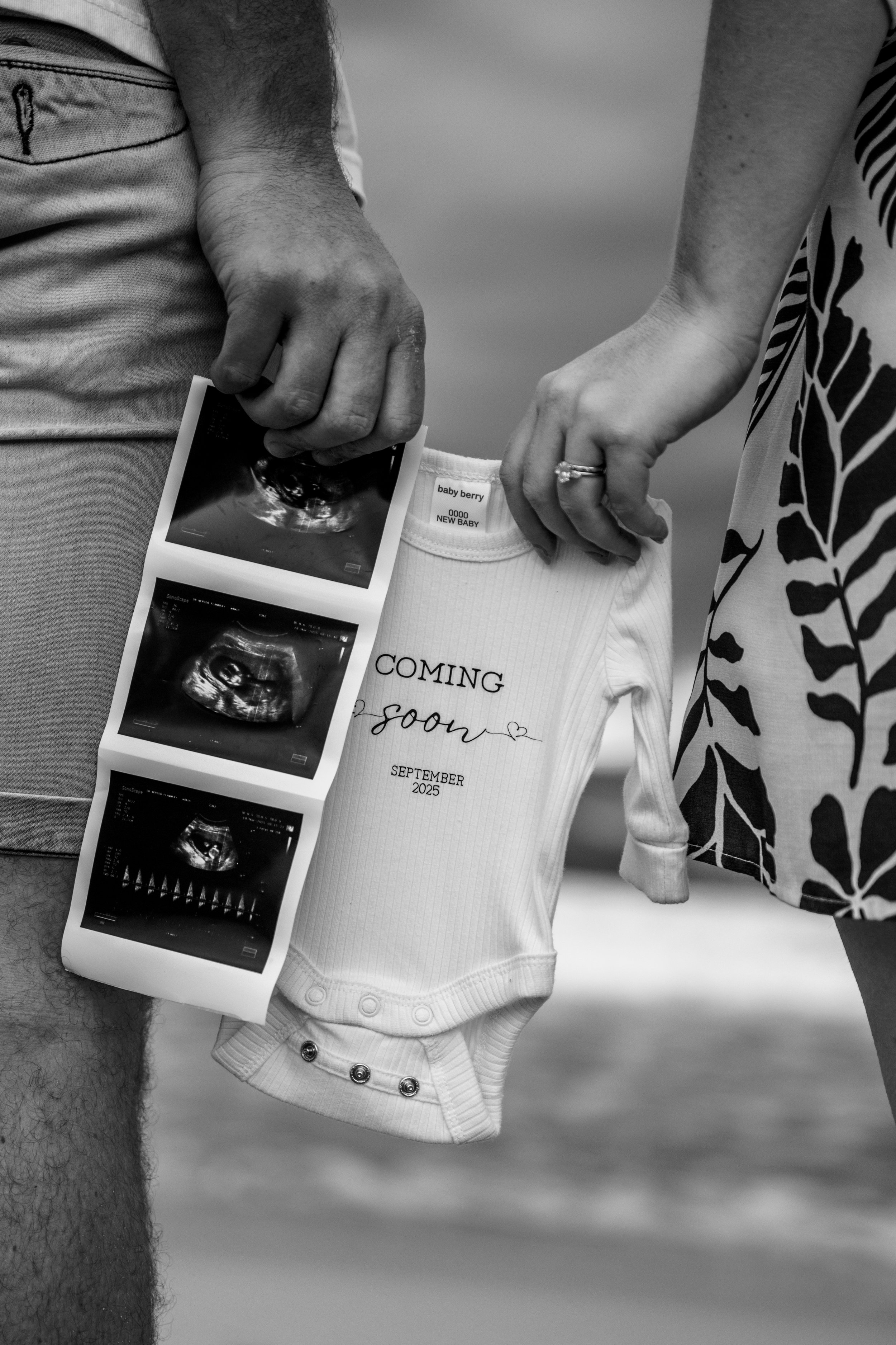 A couple at the beach holding an ultrasound printout, baby clothes, and a floating ring, indicating pregnancy announcement.