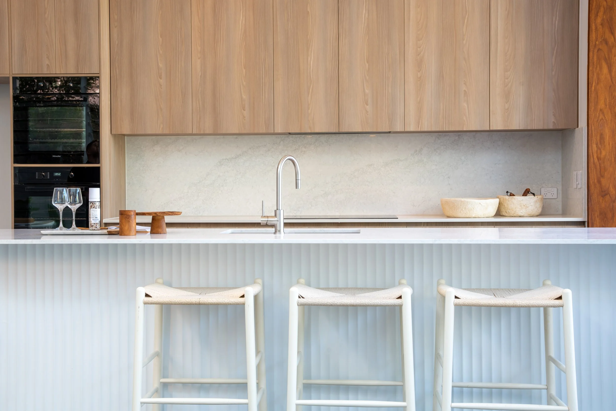 Modern kitchen with light wood cabinets, a marble backsplash, and a sleek white countertop with three bar stools in front.