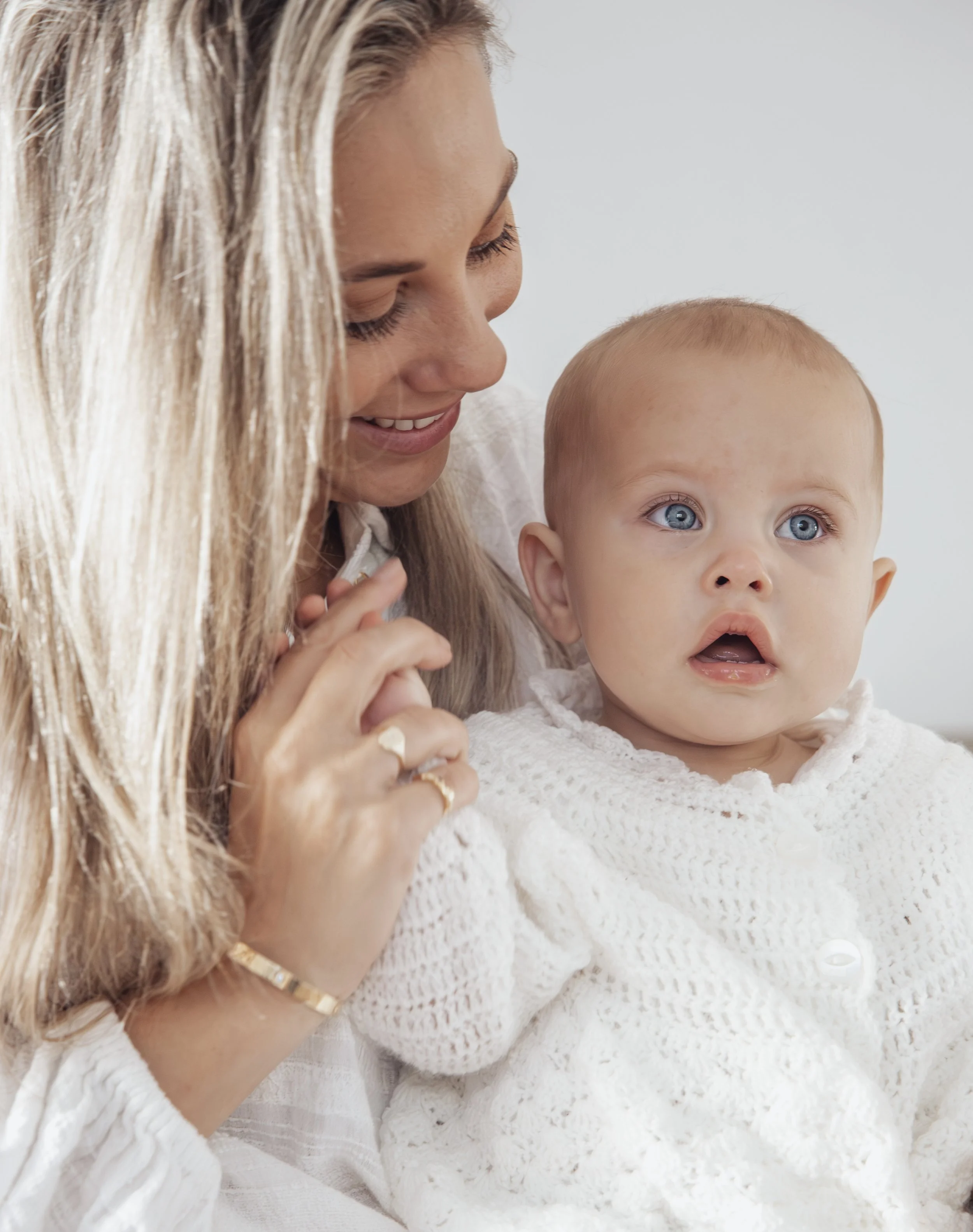 A smiling woman with long blonde hair holding a curious baby with blue eyes, both wearing white sweaters, against a plain light background.
