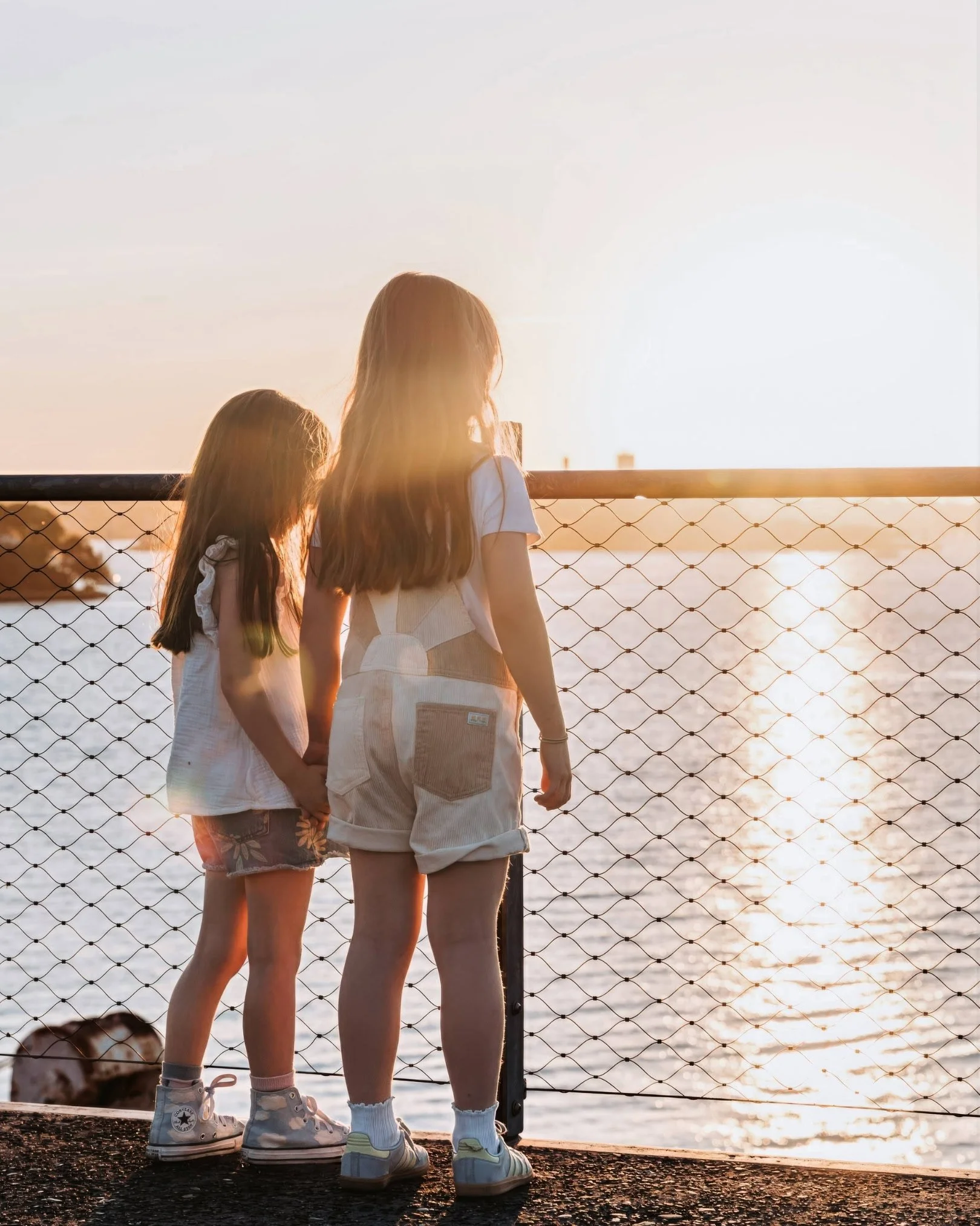 Two girls holding hands and looking at the sunset over the water near a metal railing.