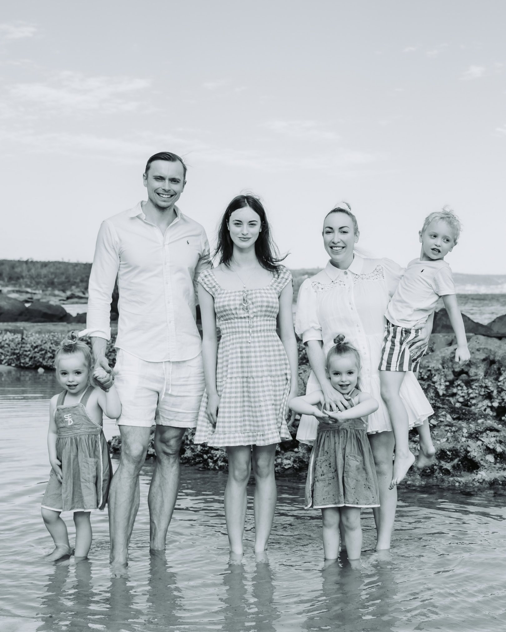 A family of seven at the beach, standing in shallow water with rocks in the background, smiling for the camera.