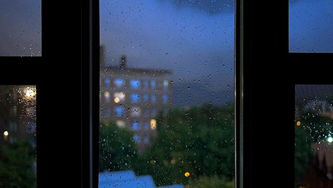 View through a rainy window at a blurry cityscape with trees and a building in the background, illuminated by city lights.