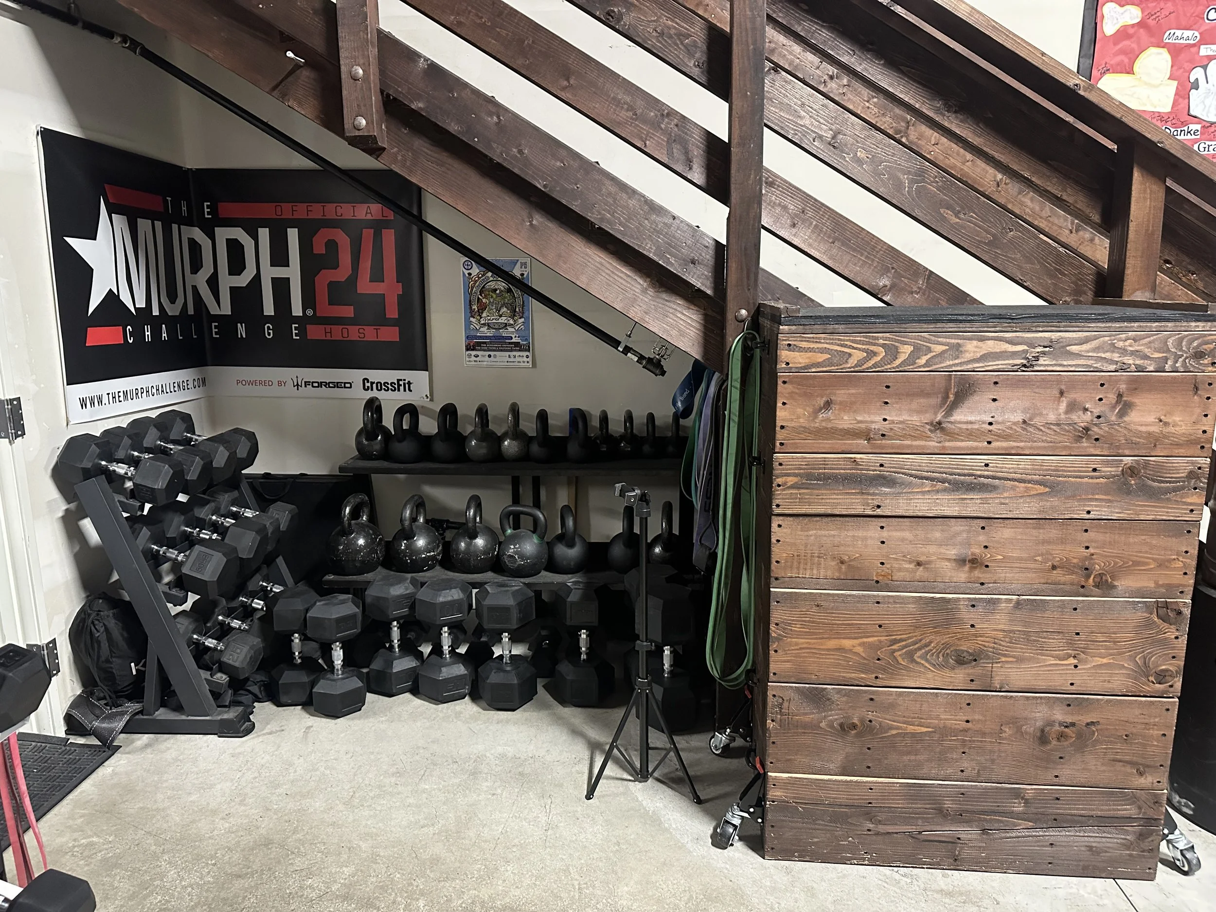 Workout area with dumbbells and kettlebells under a staircase with wooden railing, gym equipment, a banner, and resistance bands hanging.